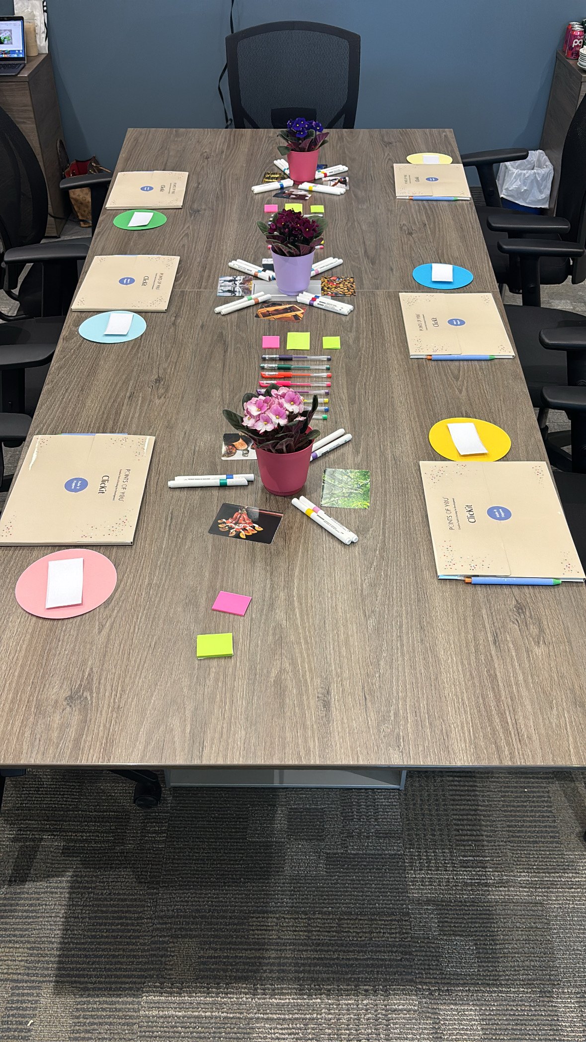 A rectangular conference table with six colored circles, each with a stack of white paper, six notebooks or folders labeled "Points of View" and "Clickit," four pots of flowering plants, and various colored sticky notes and markers arranged on top.