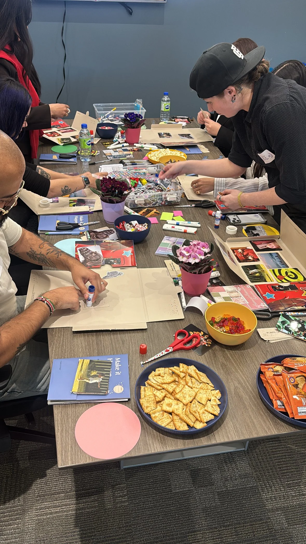 A group of people gathered around a table engaging in arts and crafts with various supplies, snacks, and drinks on the table.