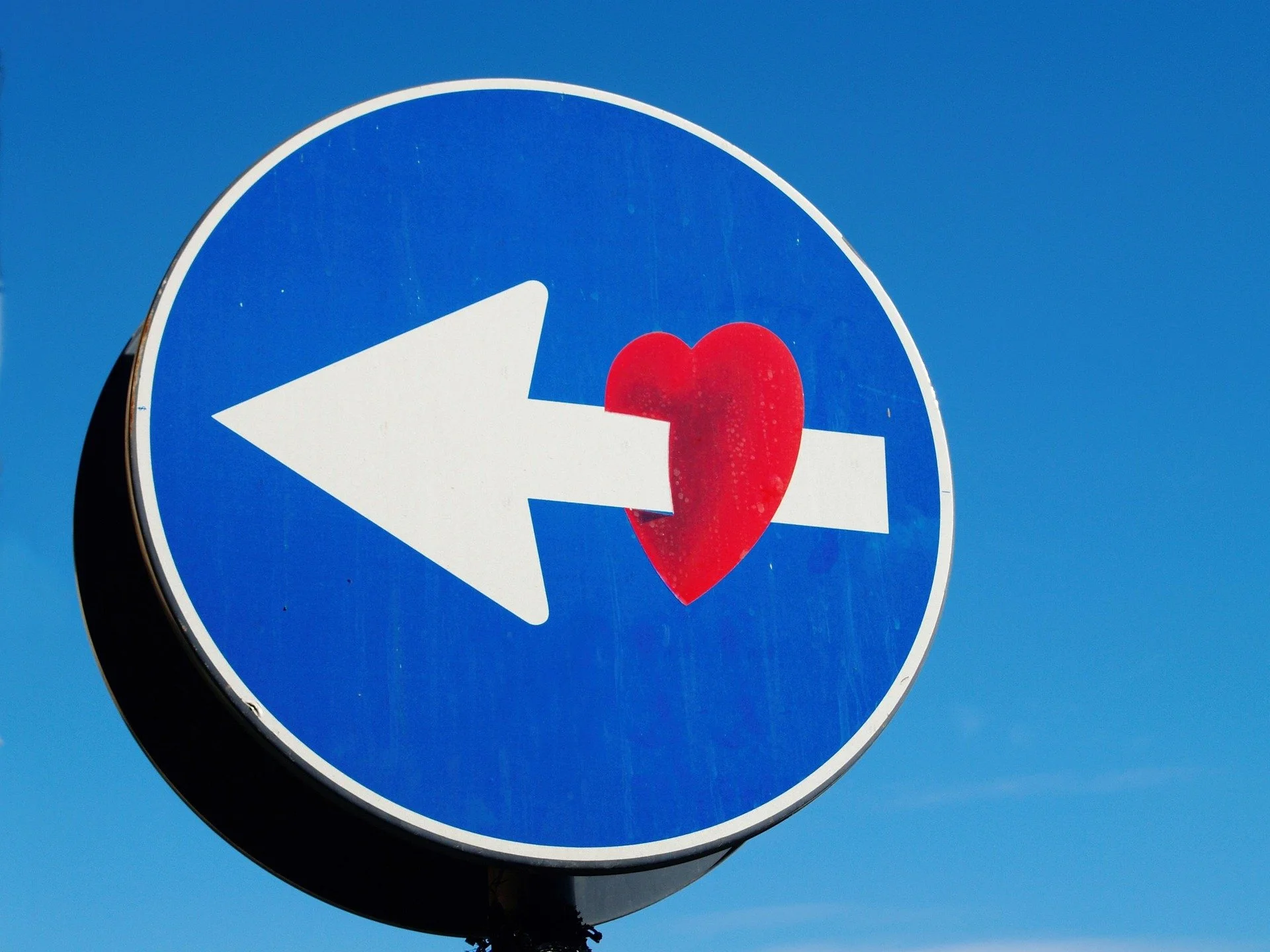 A round traffic sign with a white arrow pointing left and a red heart overlapping the arrow.