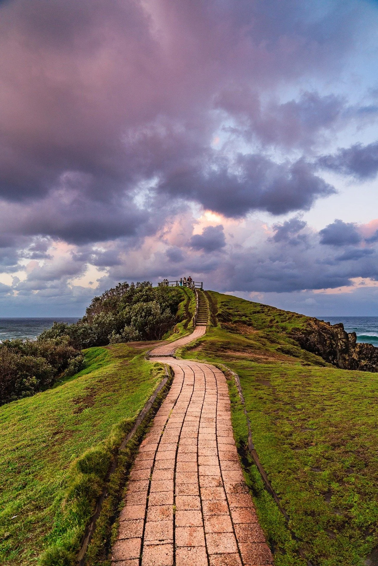 A winding brick pathway leading to a hilltop with a wooden fence under a cloudy sky at sunset.