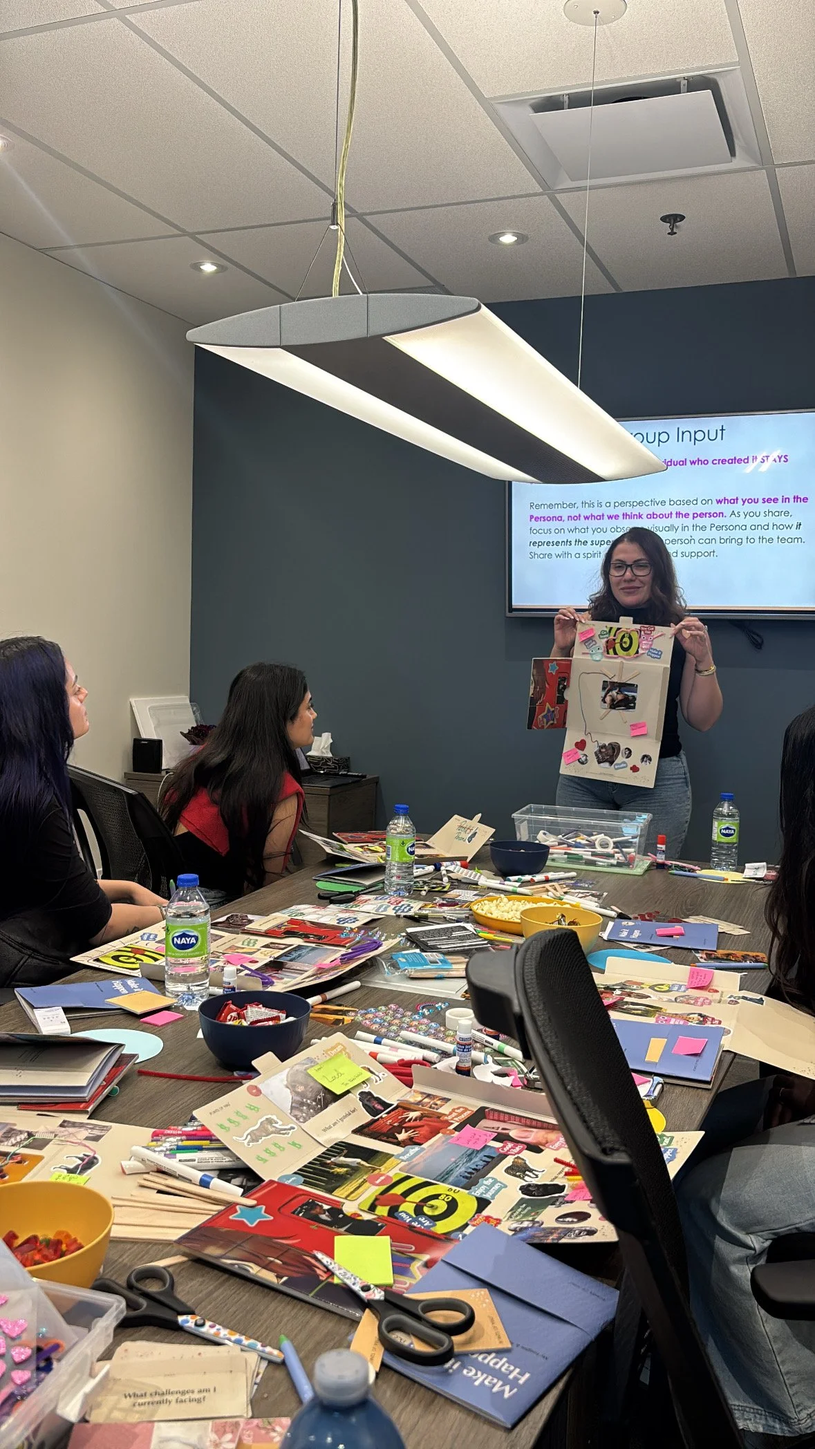A woman presenting a decorated display board to a group of seated women in a meeting room. The table is cluttered with craft supplies, notebooks, and snacks.