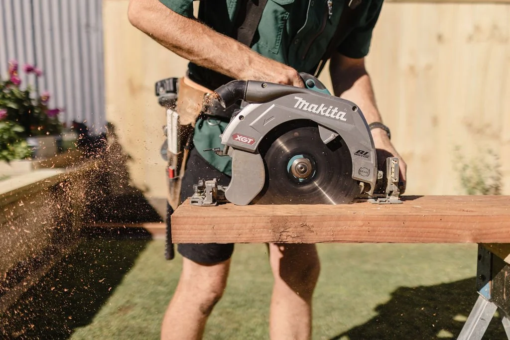 A person using a Makita circular saw to cut a wooden plank outdoors.
