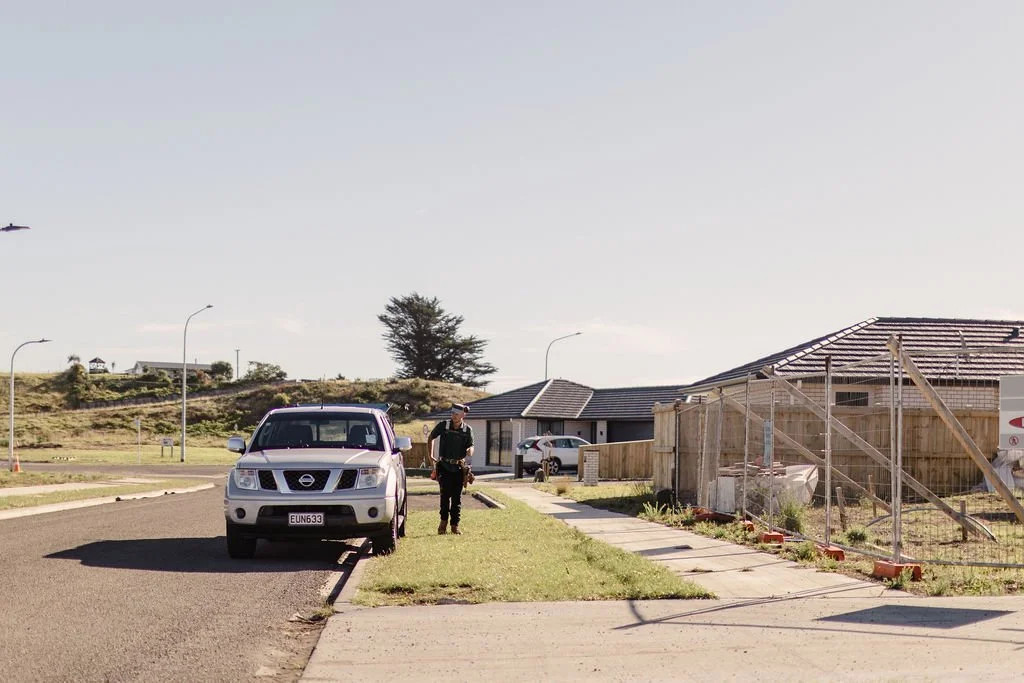 A police officer standing on the sidewalk next to a silver Nissan SUV parked on the street in a residential neighborhood under a clear sky.