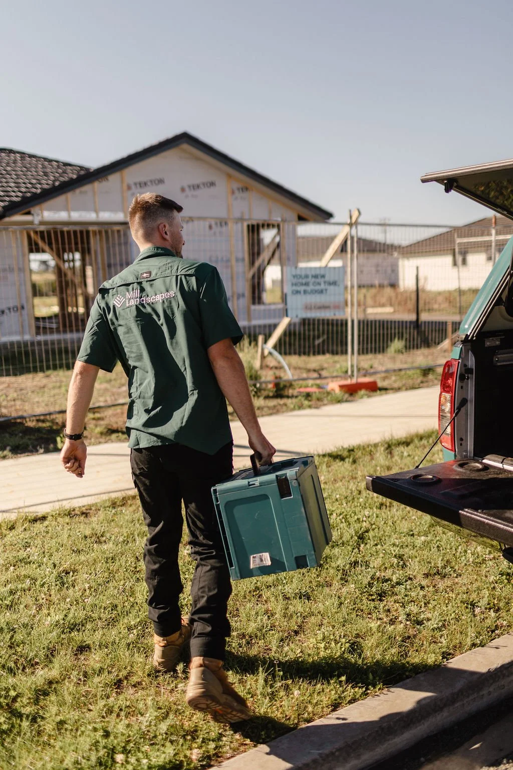 A man carrying a toolbox walks towards a house under construction, with a pickup truck nearby and a sign in the background reading "Your new home on time, on budget."