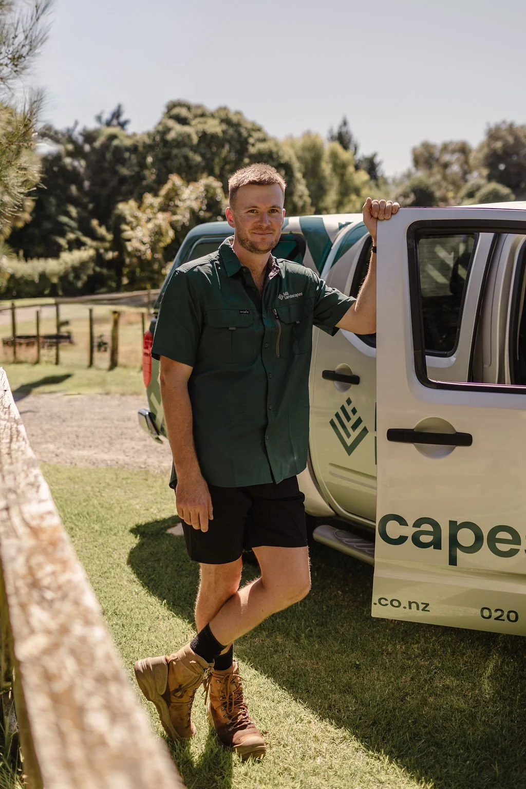 A man standing outside next to a white vehicle on a sunny day, wearing a dark green uniform shirt, black shorts, brown hiking boots, and black socks, with a background of trees and a grassy area.
