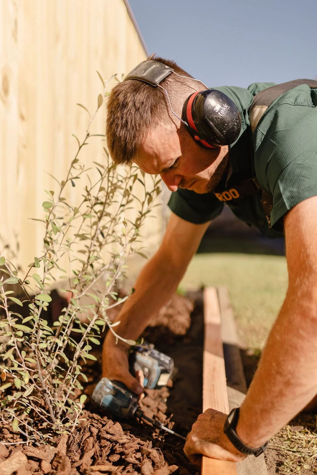 A man with brown hair wearing hearing protection, a black watch, and a green shirt is installing a wooden border along a flower bed, using a cordless drill. He is kneeling on the ground near a wooden fence, with a small plant visible nearby.