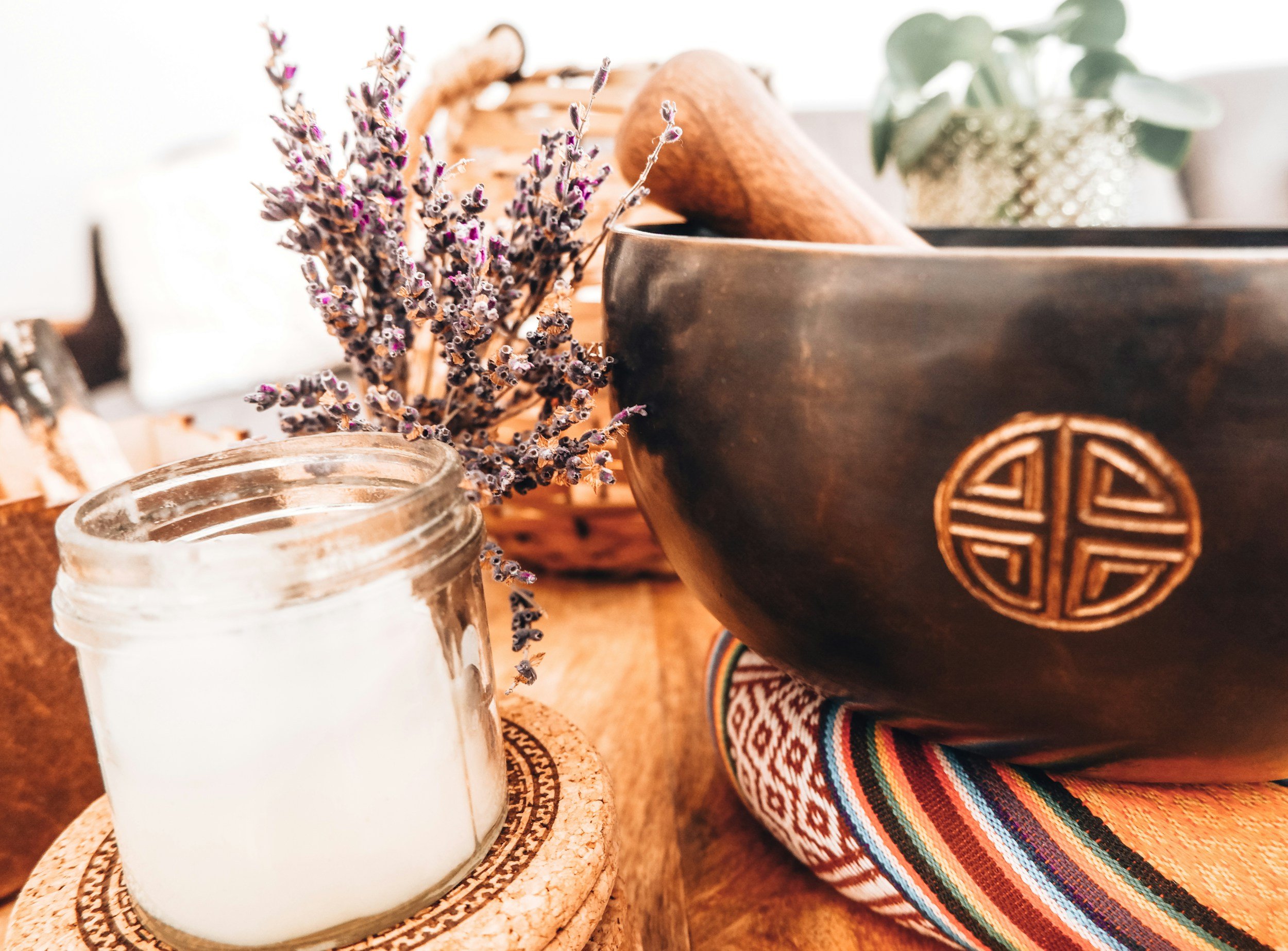 Decorative arrangement with a black singing bowl, a bunch of lavender flowers, a lit white candle in a glass jar, and some potted plants on a wooden surface with a colorful fabric underneath.