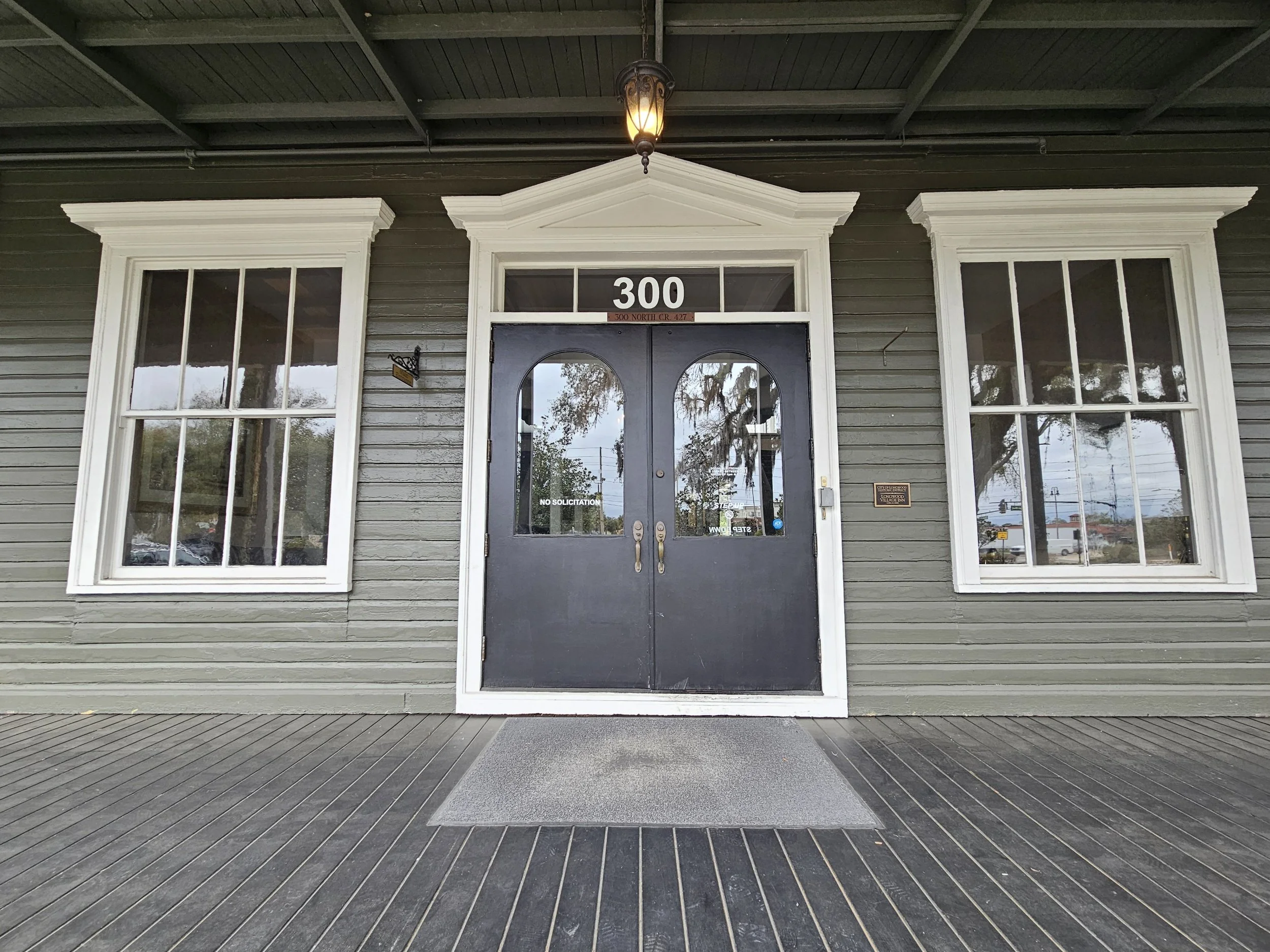 Front entrance of a building with gray walls, white window frames, black double doors with glass panels, a small lantern hanging above, and a gray welcome mat on a wooden porch.