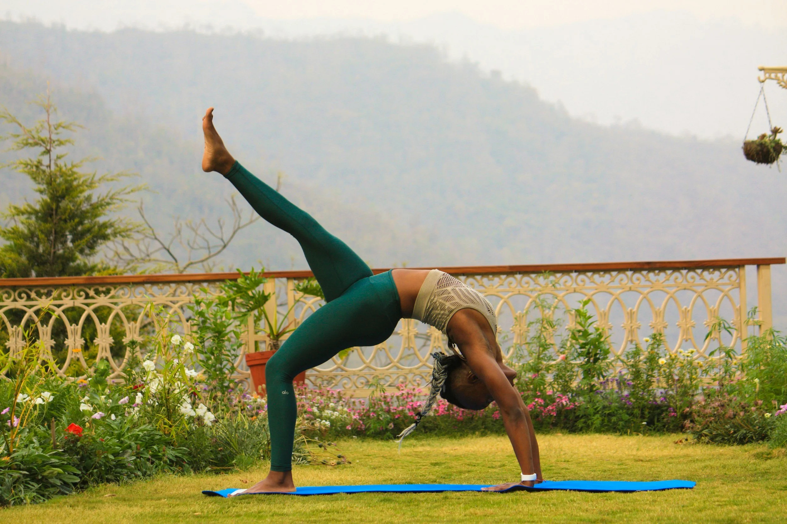 A woman practicing yoga outdoors on a blue mat, performing a downward dog pose with one leg raised, surrounded by plants and flowers with a decorative railing and foggy mountains in the background.