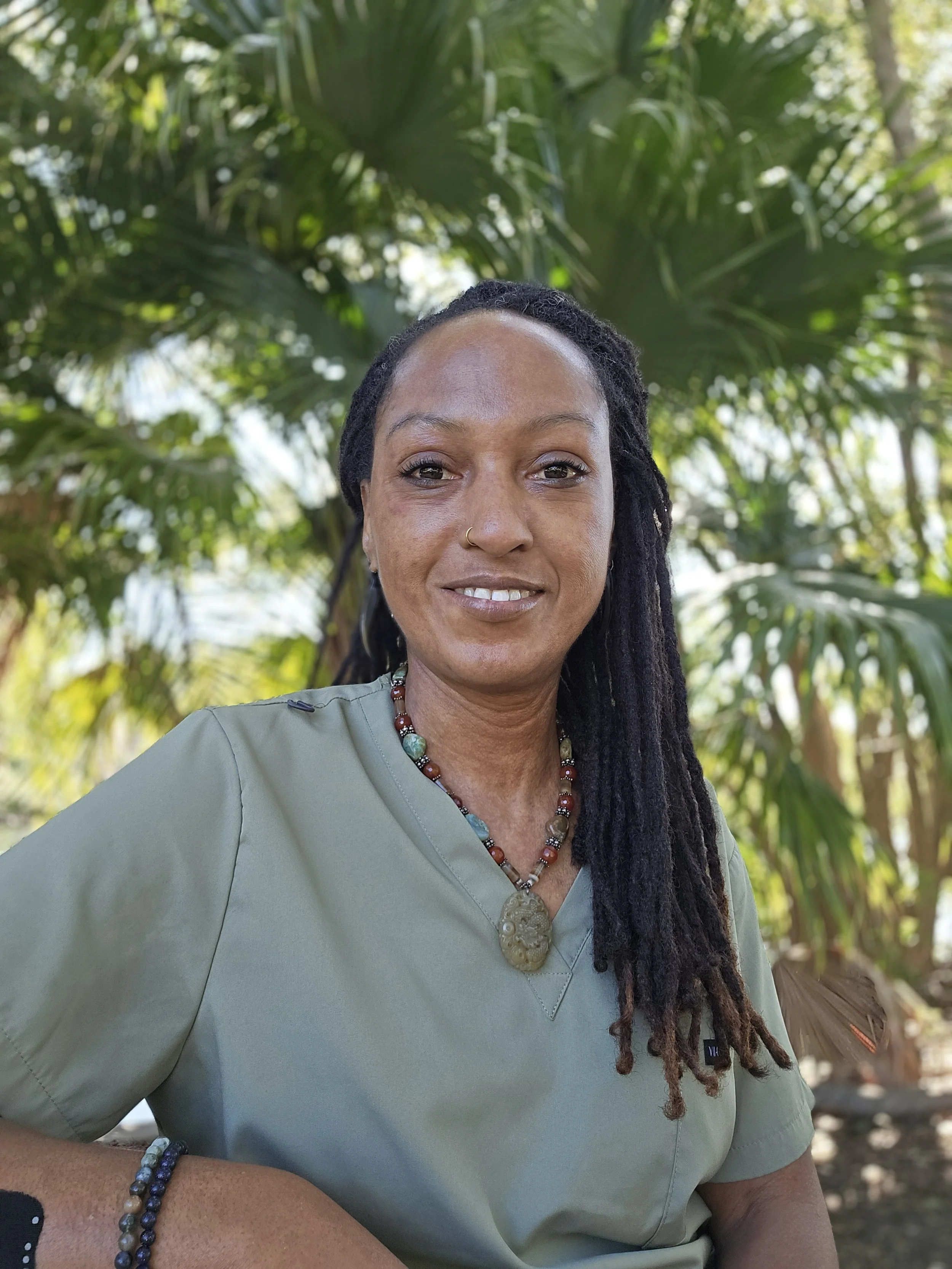 A person practicing yoga in a seated meditation pose on a wooden floor at sunrise or sunset with palm trees in the background.