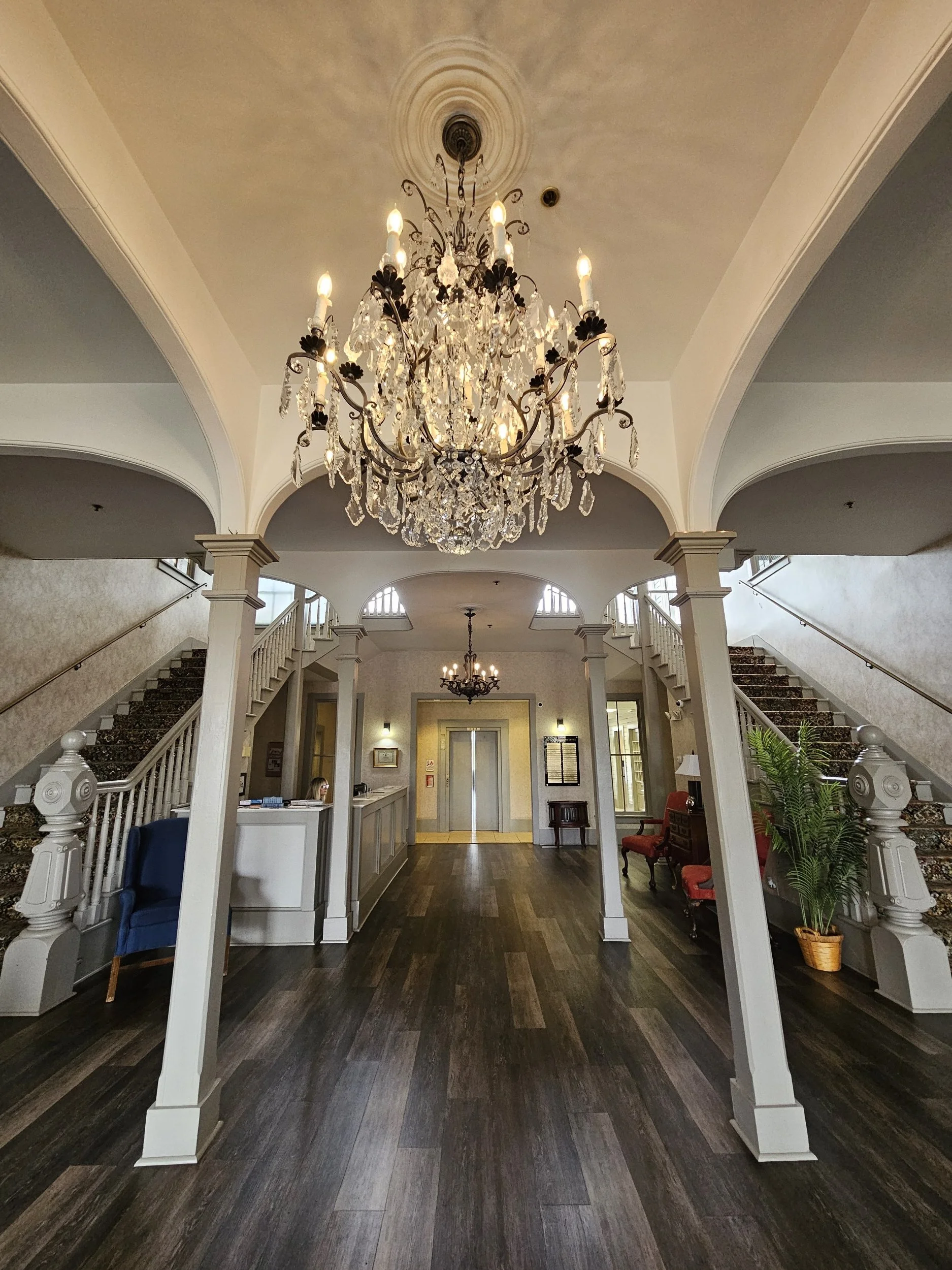 Elegant hotel lobby with a large crystal chandelier hanging from the ceiling, a reception desk, a staircase on each side, and a seating area with chairs and a potted plant.