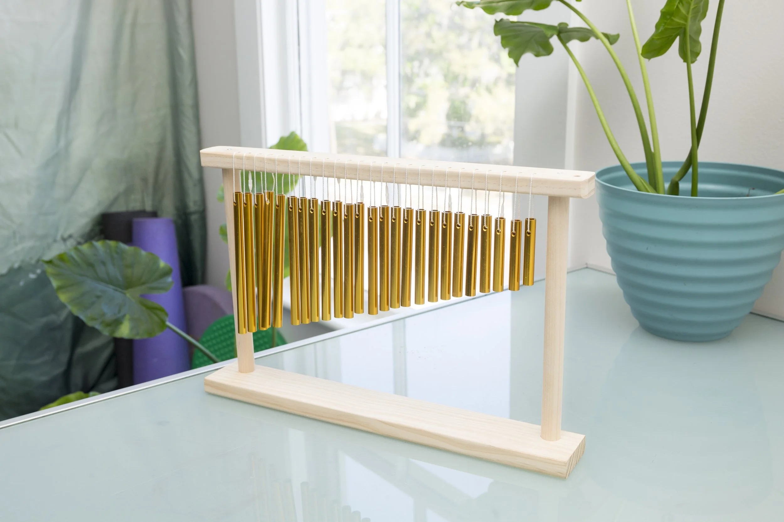 A wooden chime with gold metal tubes hanging from a white frame, placed on a white table next to a large green indoor plant in a turquoise pot, with a window in the background.