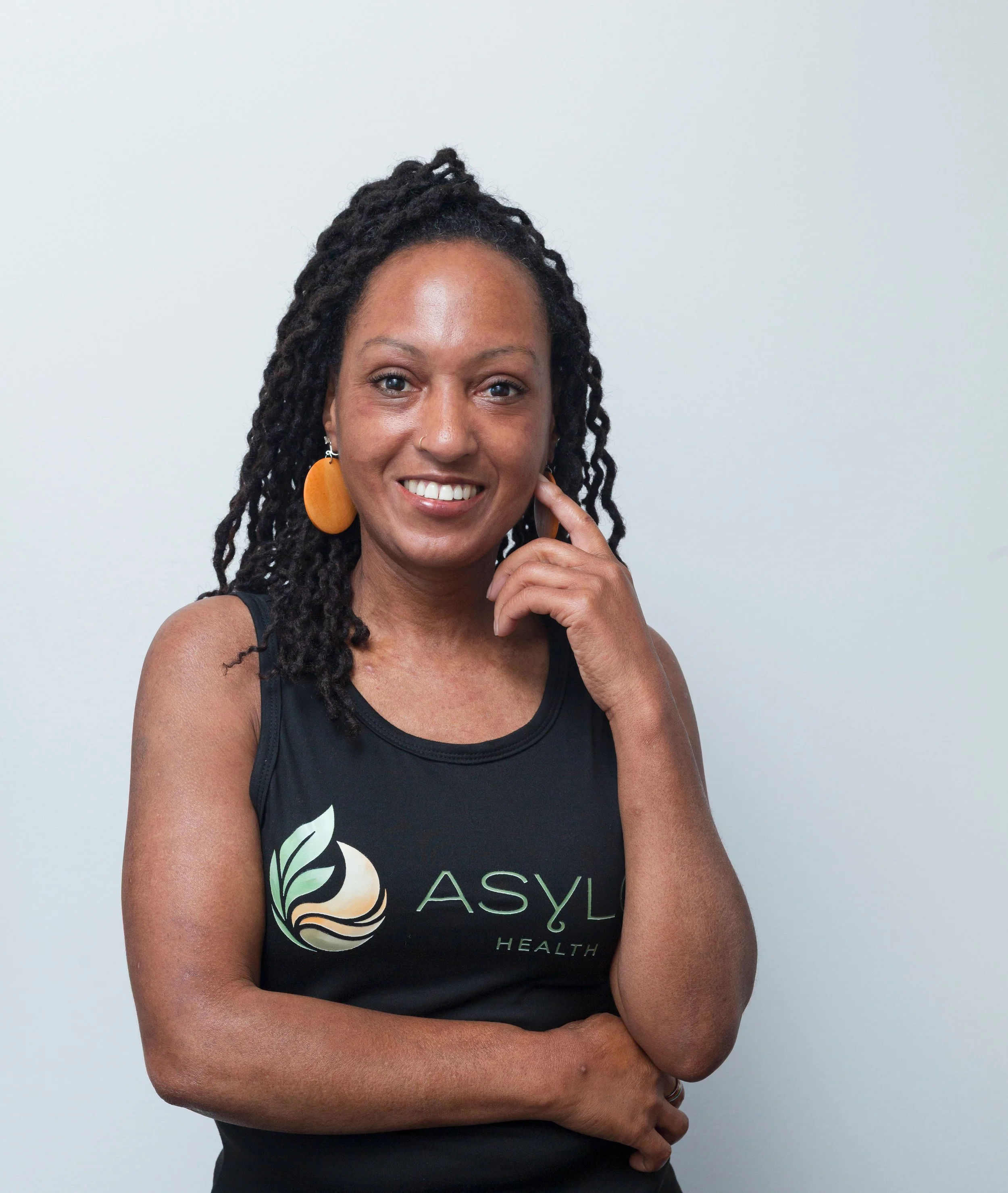 A woman with locs smilinglarge earrings, and a black tank top with the Asylo Health logo, standing against a plain light gray wall.