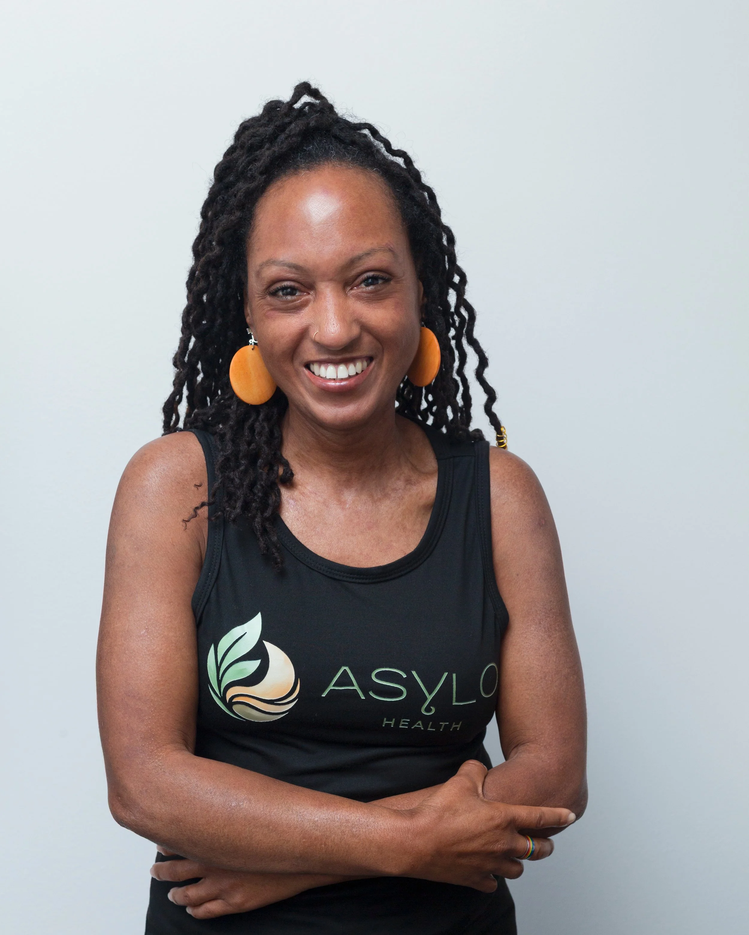 A woman with locs wearing large orange earrings and a black tank top with a logo that says ASYLO Health, standing against a plain white background, smiling and crossing her arms.