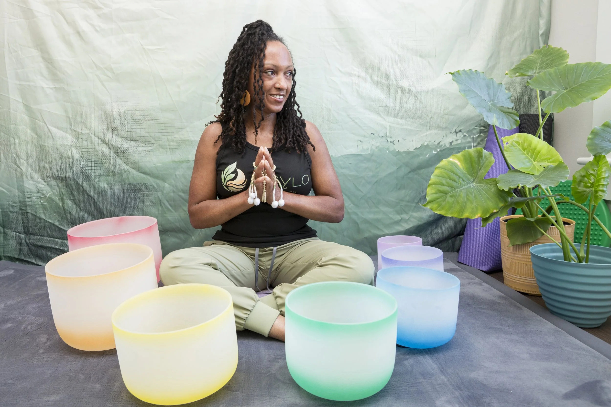 A woman with locs, wearing a black tank top and beige pants, sitting cross-legged on the floor with her hands in a prayer position, surrounded by colorful candle holders and potted plants in an indoor setting.