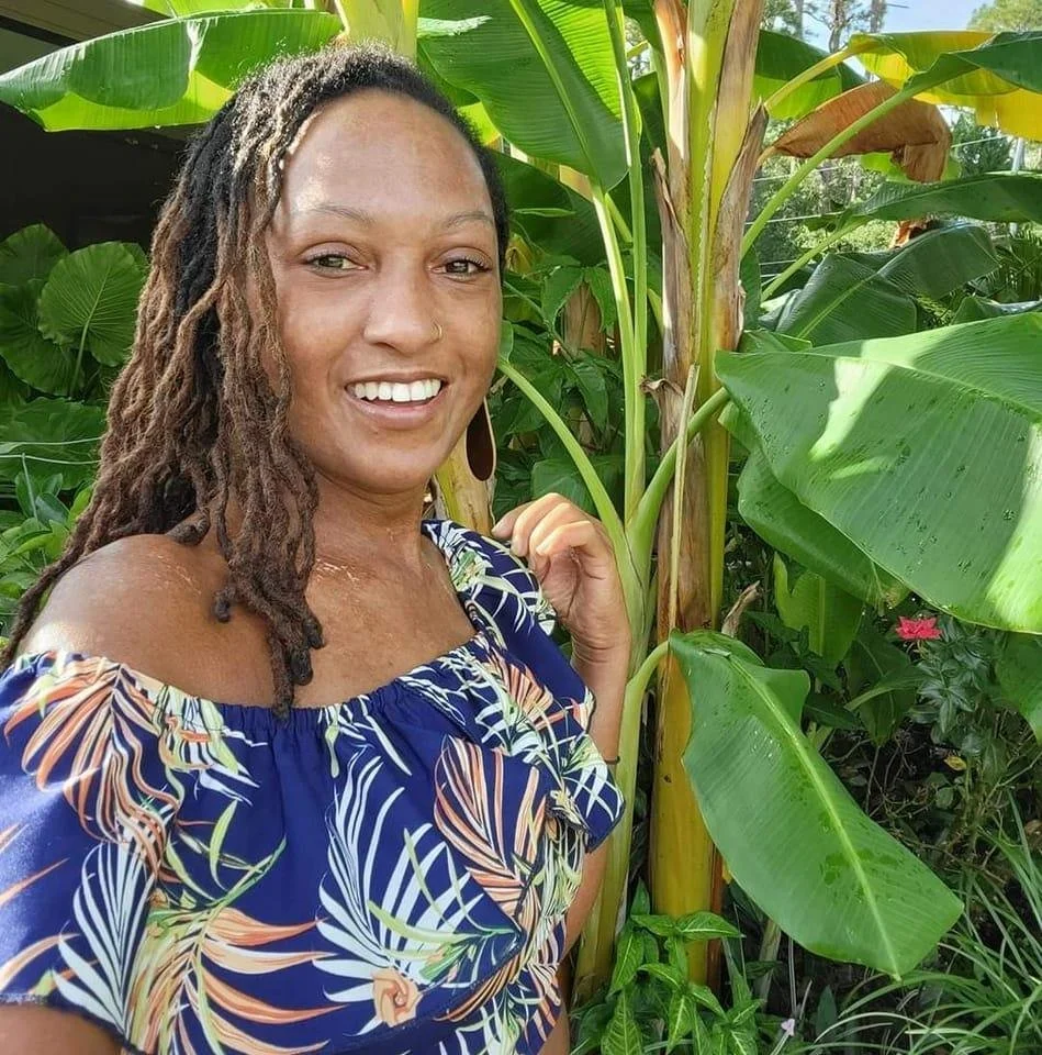 A woman with dreadlocks and a nose ring, smiling, standing among lush green banana plants in a tropical garden.