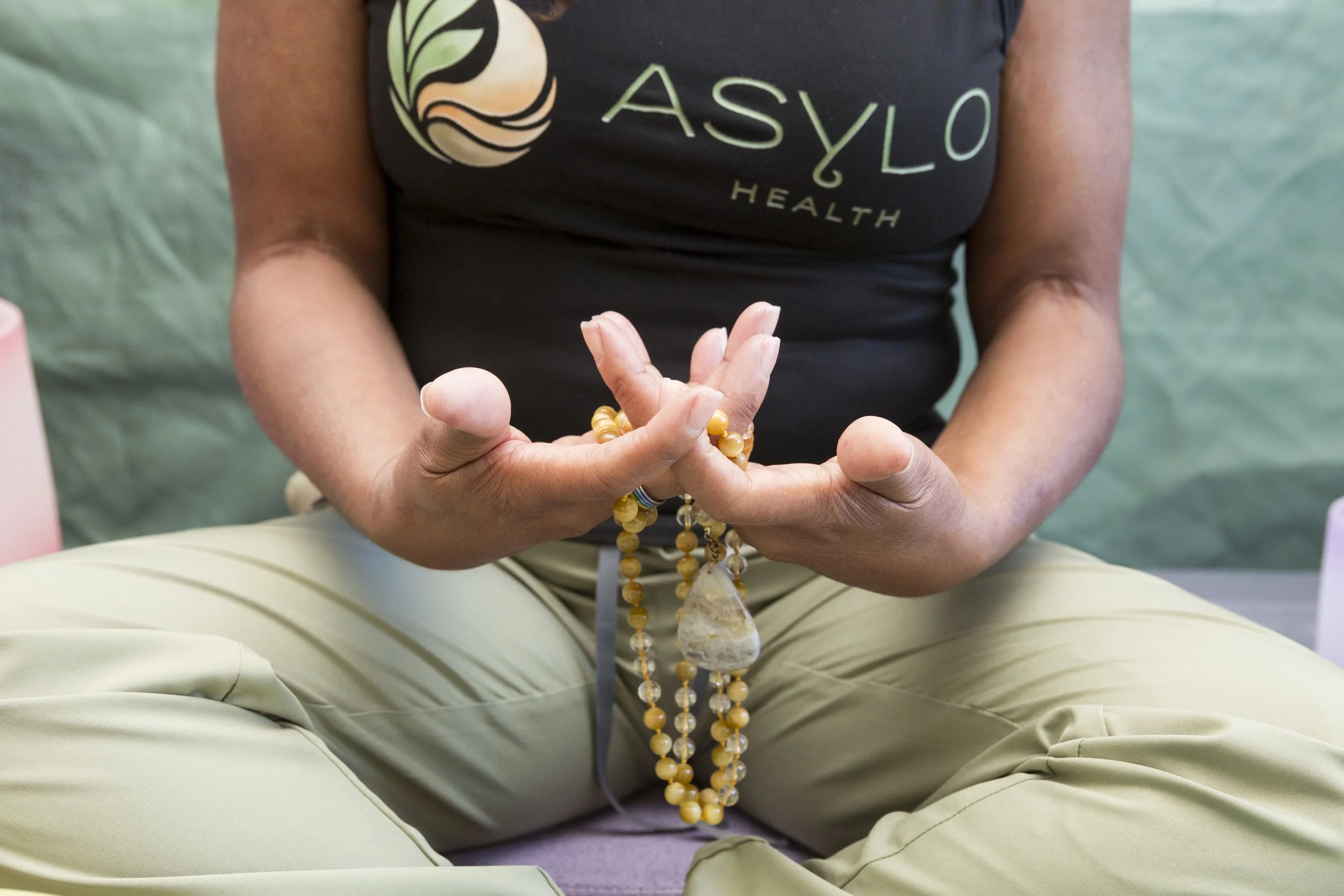 Person sitting cross-legged holding prayer beads in their hands. They are wearing a black shirt with a logo and text reading 'ASYOLO HEALTH' and beige pants. The background is a soft, out-of-focus greenish color.