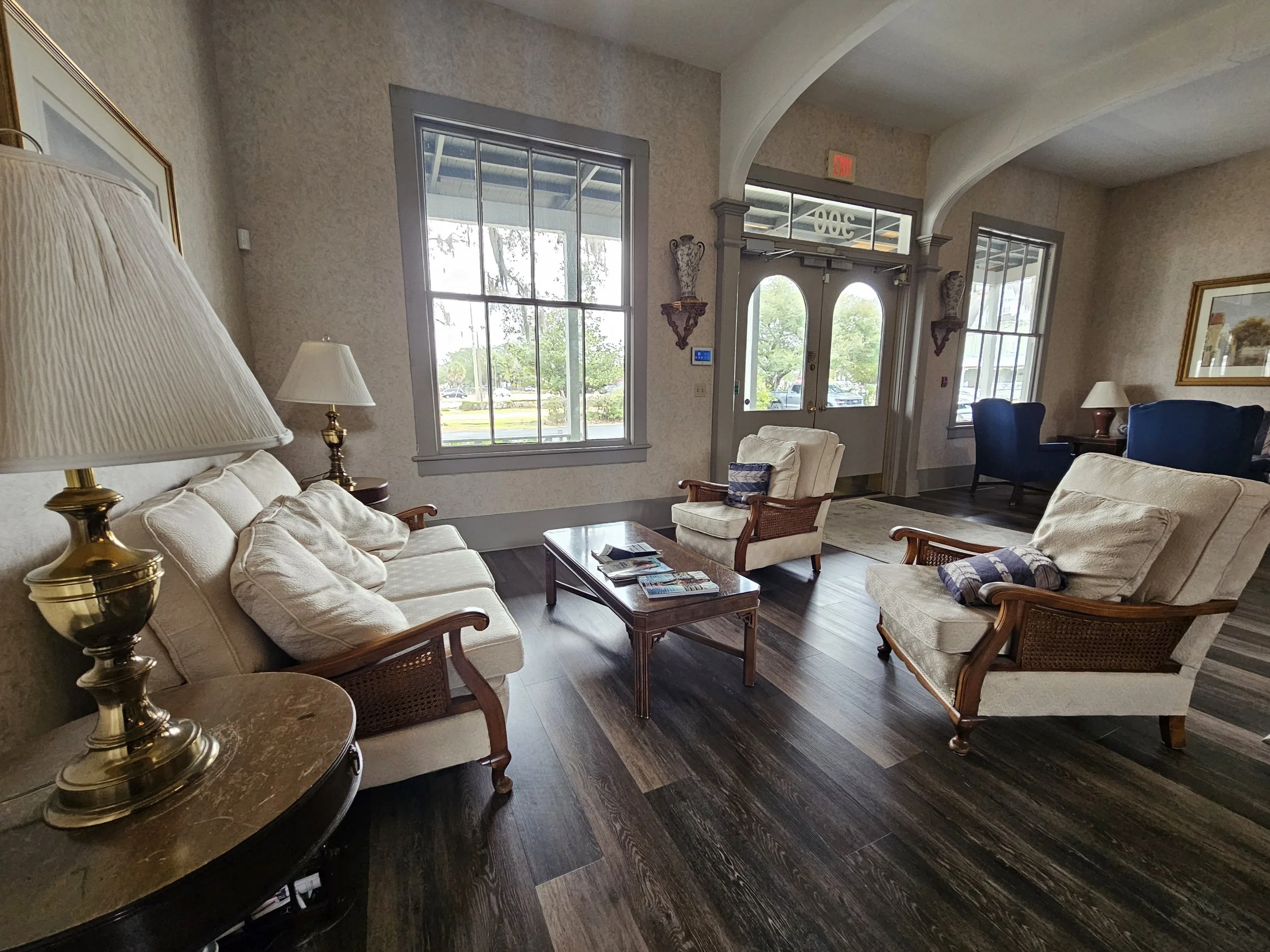 A cozy lobby area with vintage-style furniture including four armchairs and a sofa, a wooden coffee table, and table lamps. Large windows and a glass door allow natural light to brighten the space, with a view of trees and cars outside.