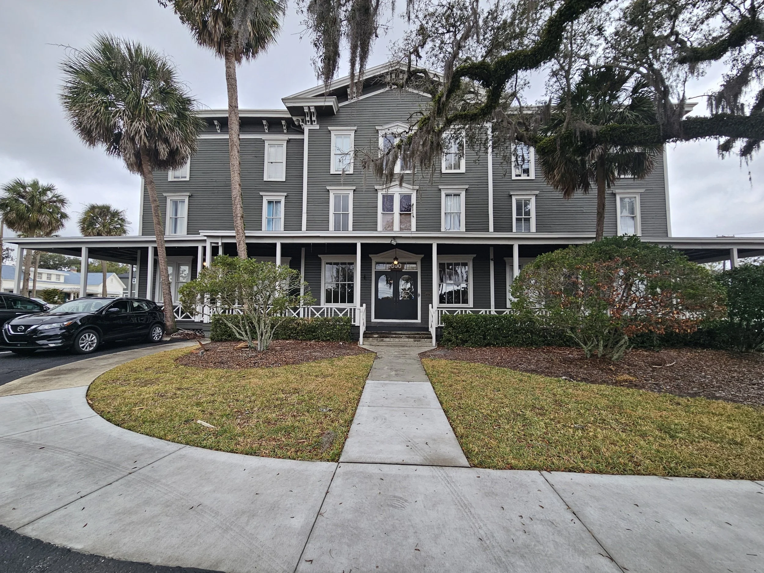 A three-story gray wooden building with white trim, set among palm trees and bushes, with a sidewalk leading to the black front door.