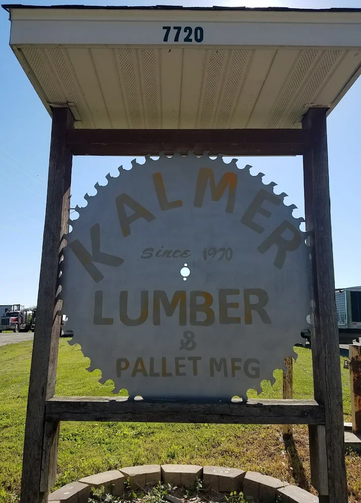 Sign for Kalmer Lumber and Pallet Manufacturing, established in 1970, with a circular saw blade design, located outdoors with a grassy area and trucks in the background.