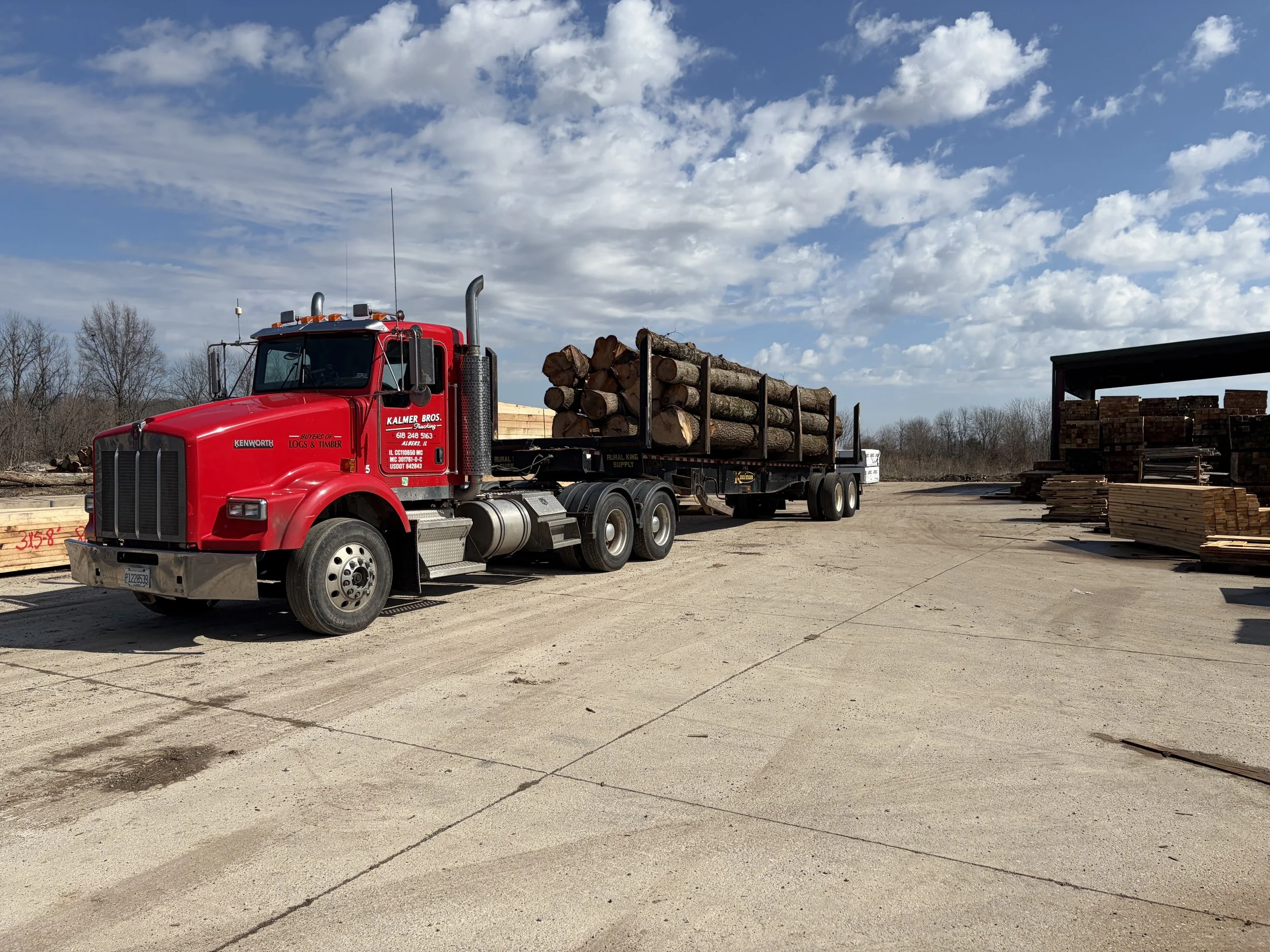 A red truck carrying cut logs is parked in an industrial outdoor lumber yard with stacks of wood and lumber on shelves, under a partly cloudy sky.