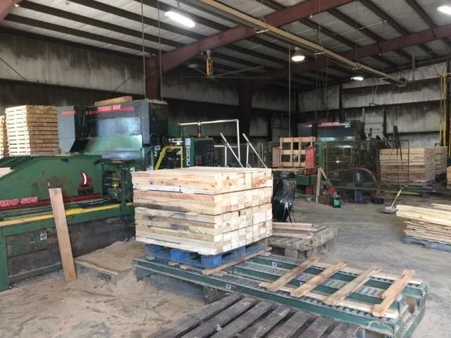 Stacks of wooden planks and pallets inside a woodworking factory or warehouse, with machinery and equipment for cutting and processing wood.