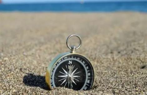 A black compass with a silver ring on sandy beach with the ocean in the background.