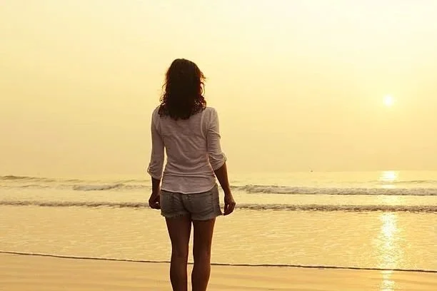 A woman standing on the beach, facing the ocean and watching the sunset.