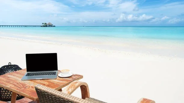 Laptop, coffee cup, and backpack on a wooden table at the beach with white sand, turquoise water, and a pier in the distance.