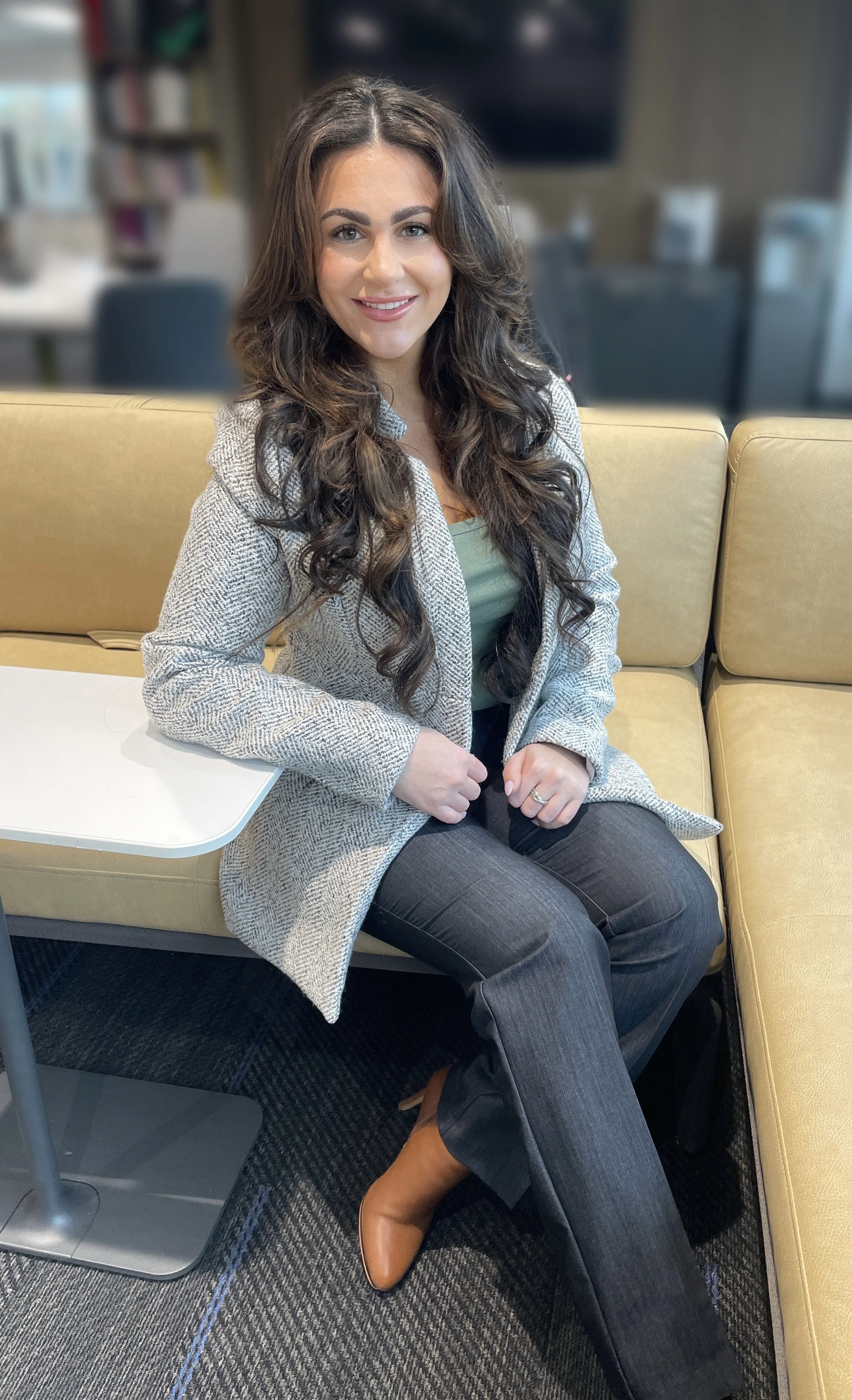 A woman with long, curly brown hair smiling, sitting on a yellow couch in a modern office space, wearing a patterned blazer, green top, black pants, and brown ankle boots.