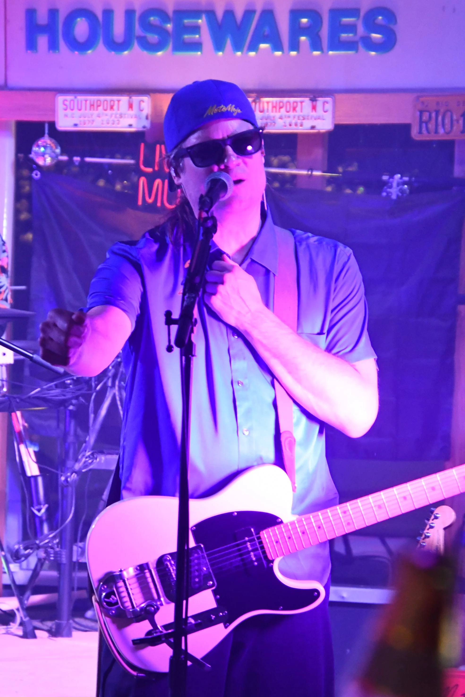 A musician on stage singing into a microphone, wearing sunglasses, a cap, and playing an electric guitar.