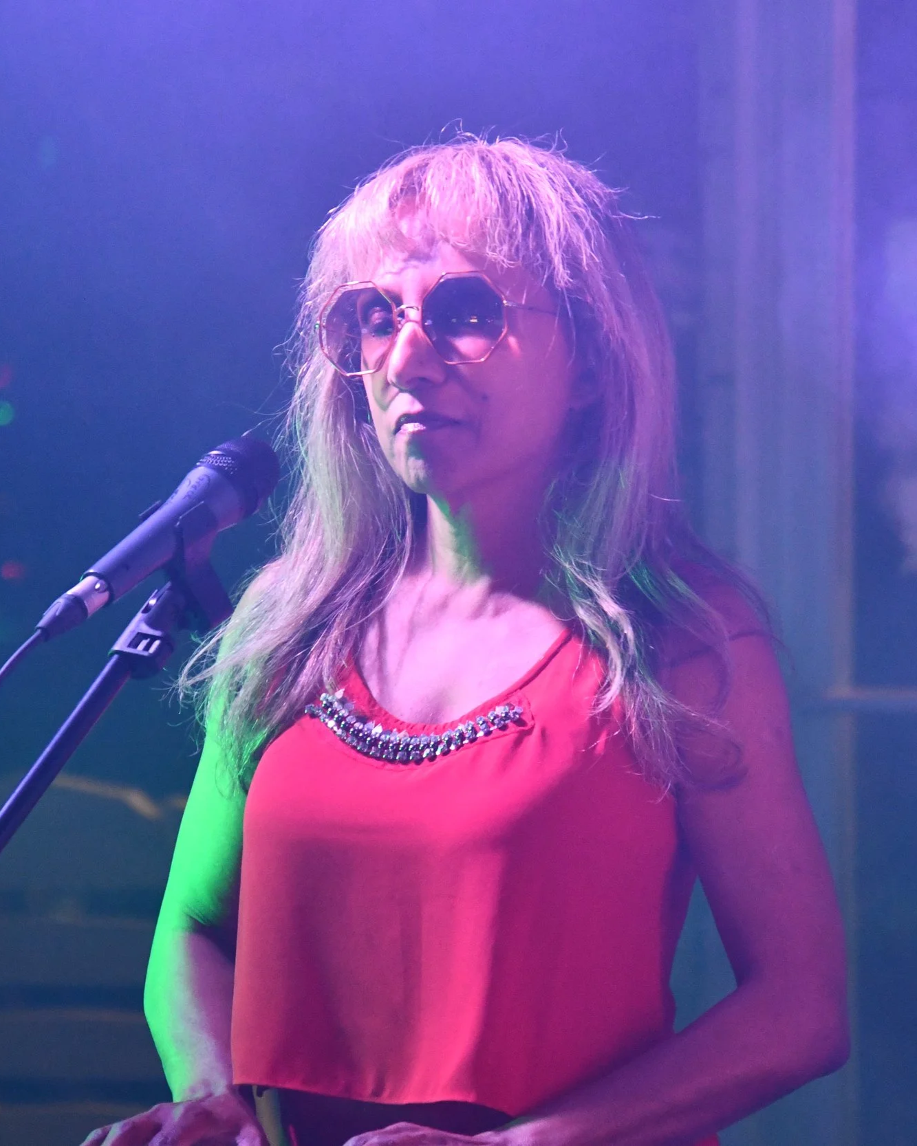 A woman with blonde hair, wearing sunglasses and a red top with a decorative neckline, standing on stage behind a microphone under purple and green lighting.