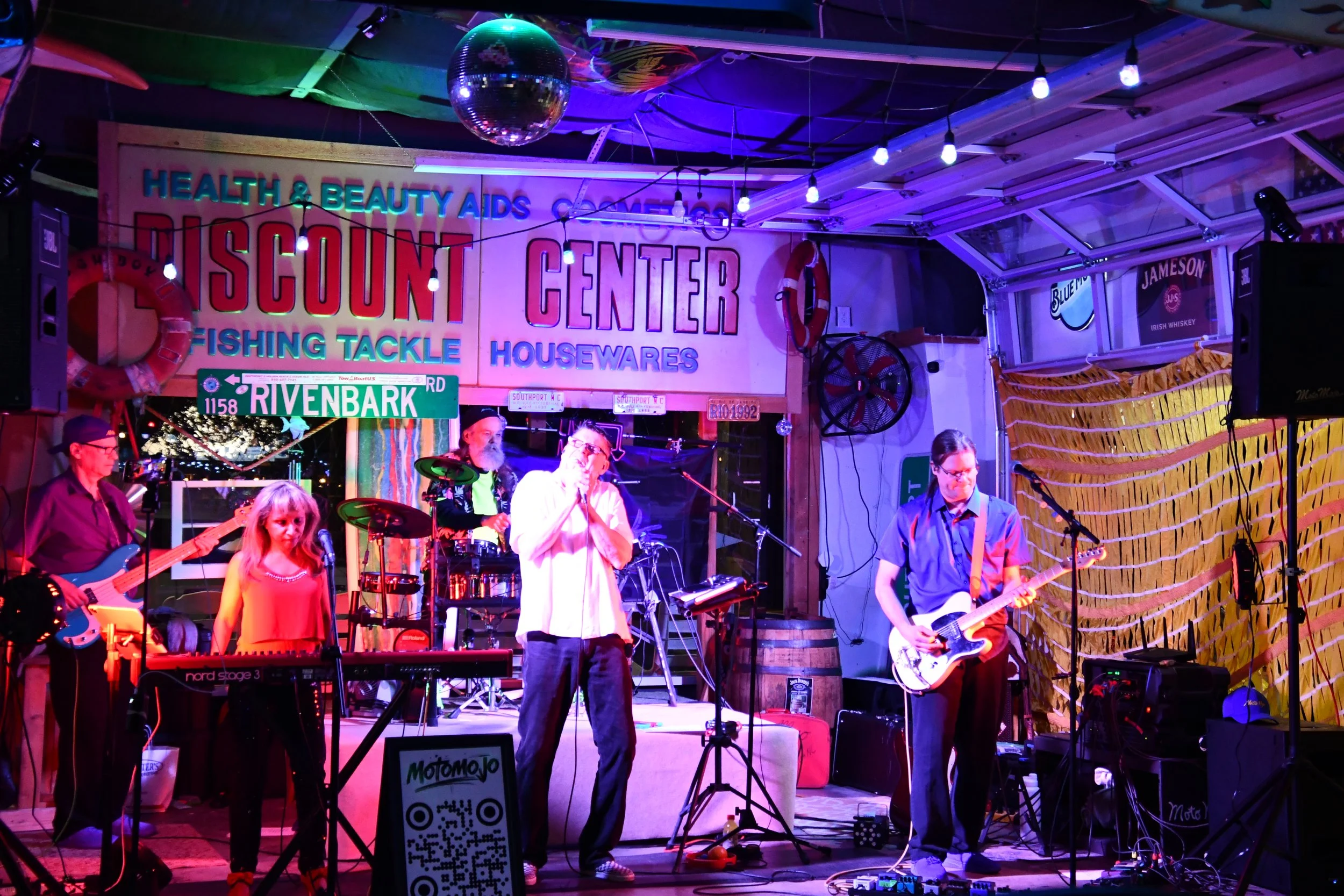 MotoMojo performing live on stage, with musical instruments and equipment visible, and colorful stage lighting.