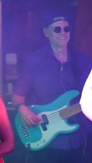 Person wearing sunglasses playing a bass guitar in a dark, colorful setting.