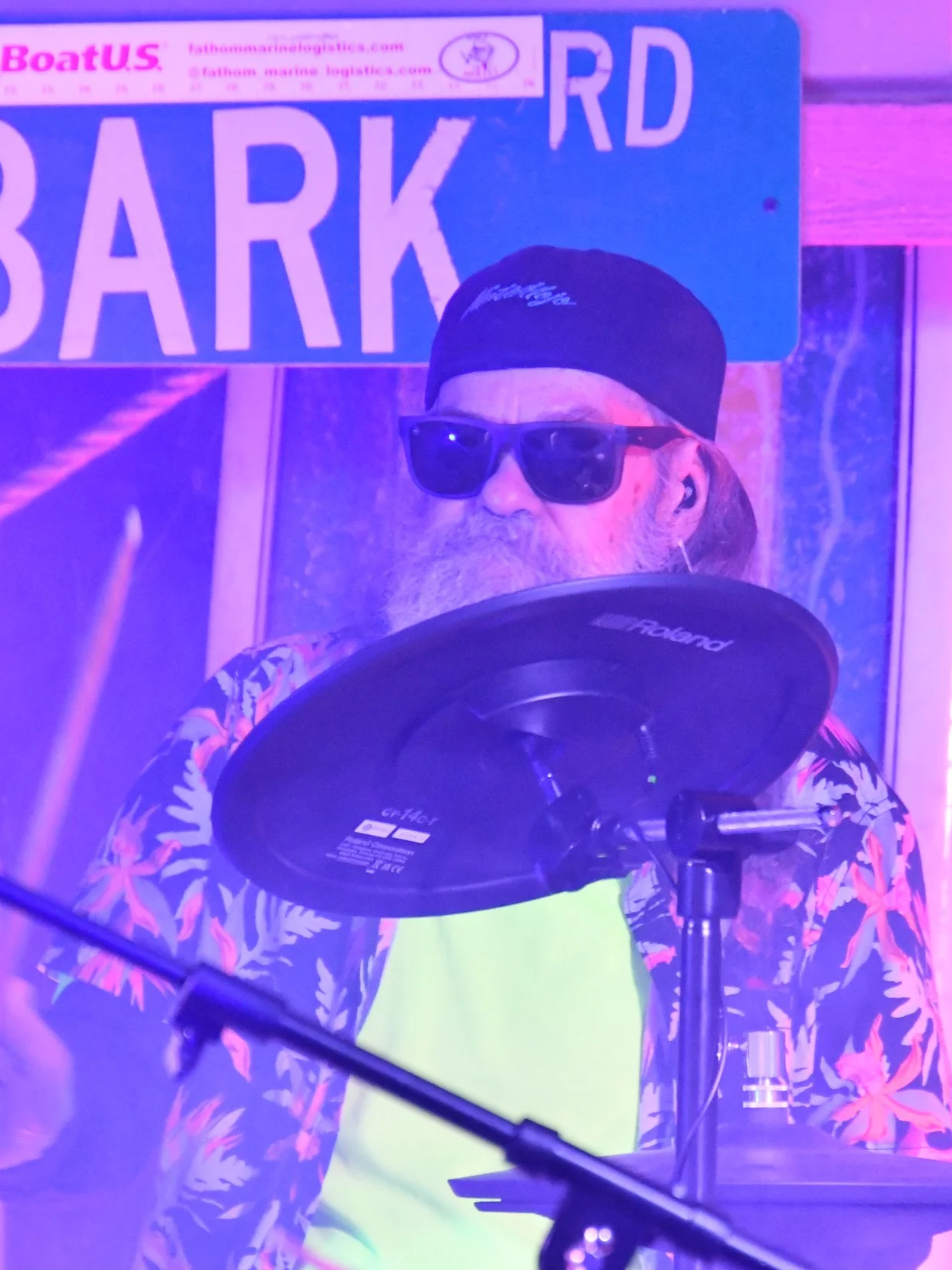 A man with a beard and sunglasses wearing a black cap and tropical shirt playing the drums.