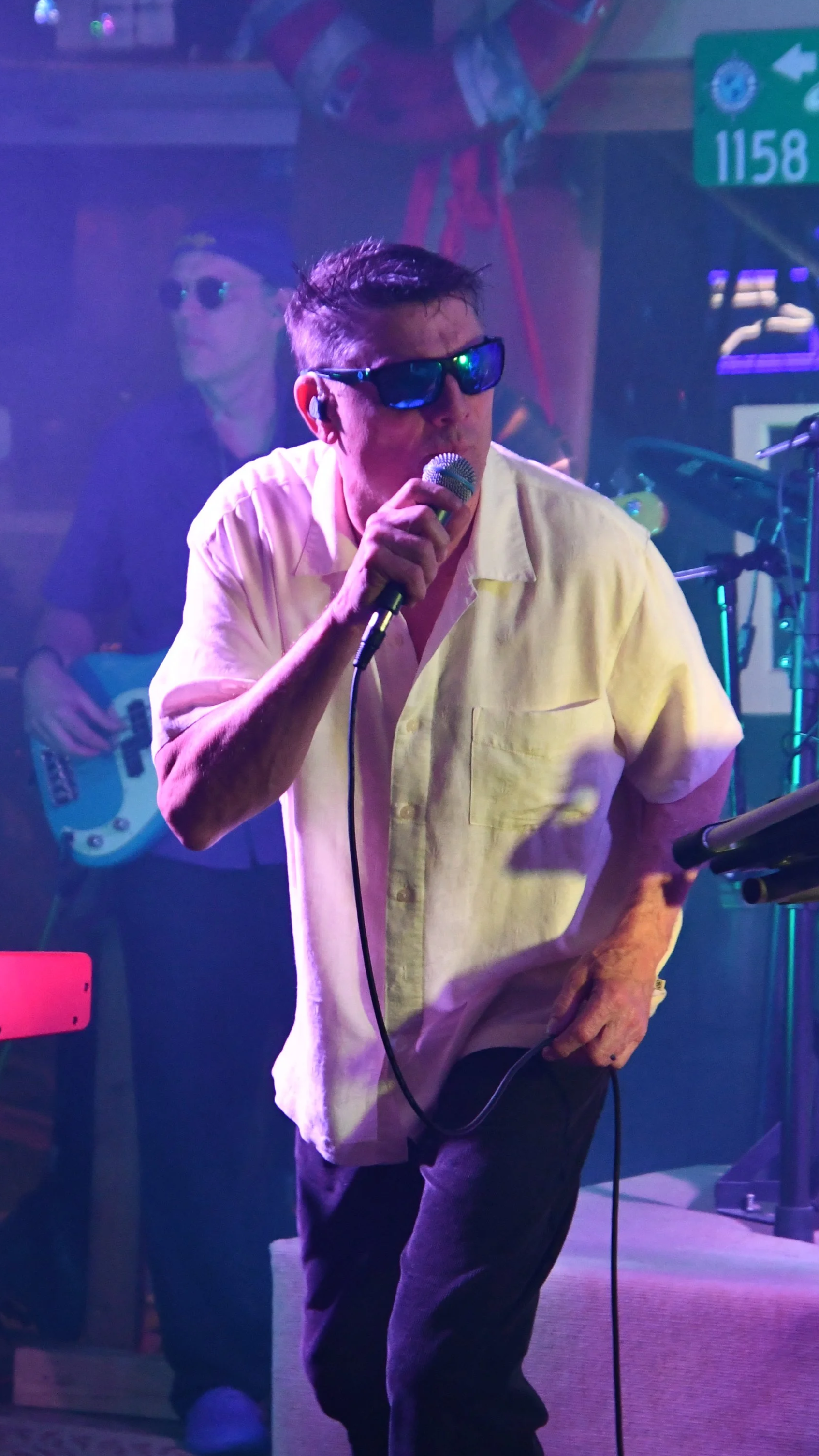 A man wearing sunglasses and a cream-colored shirt is singing into a microphone, with another musician playing guitar in the background, on a stage with colorful lighting.