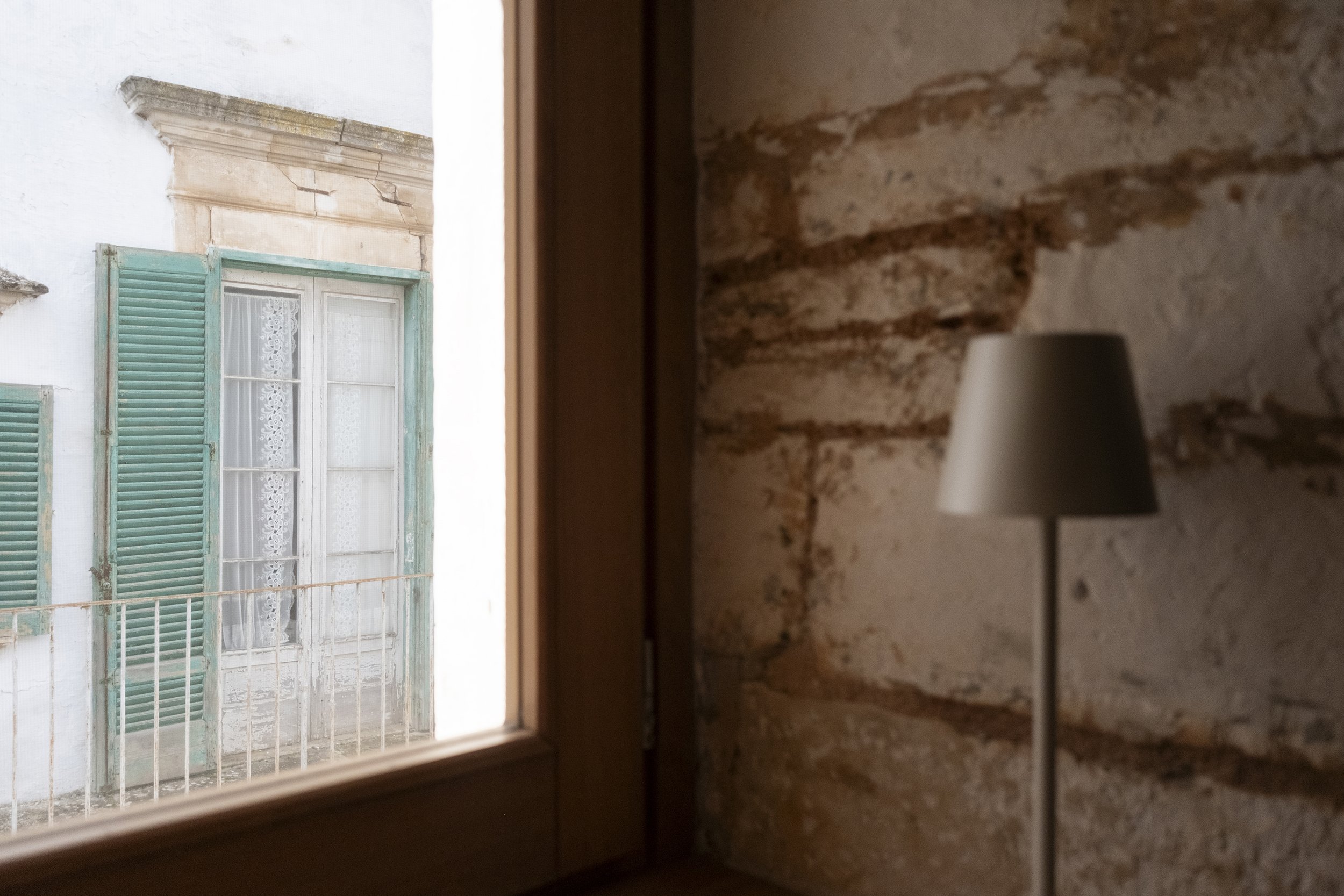 A view of an old building with a window featuring turquoise shutters, seen from inside a room with a brick wall and a white floor lamp.