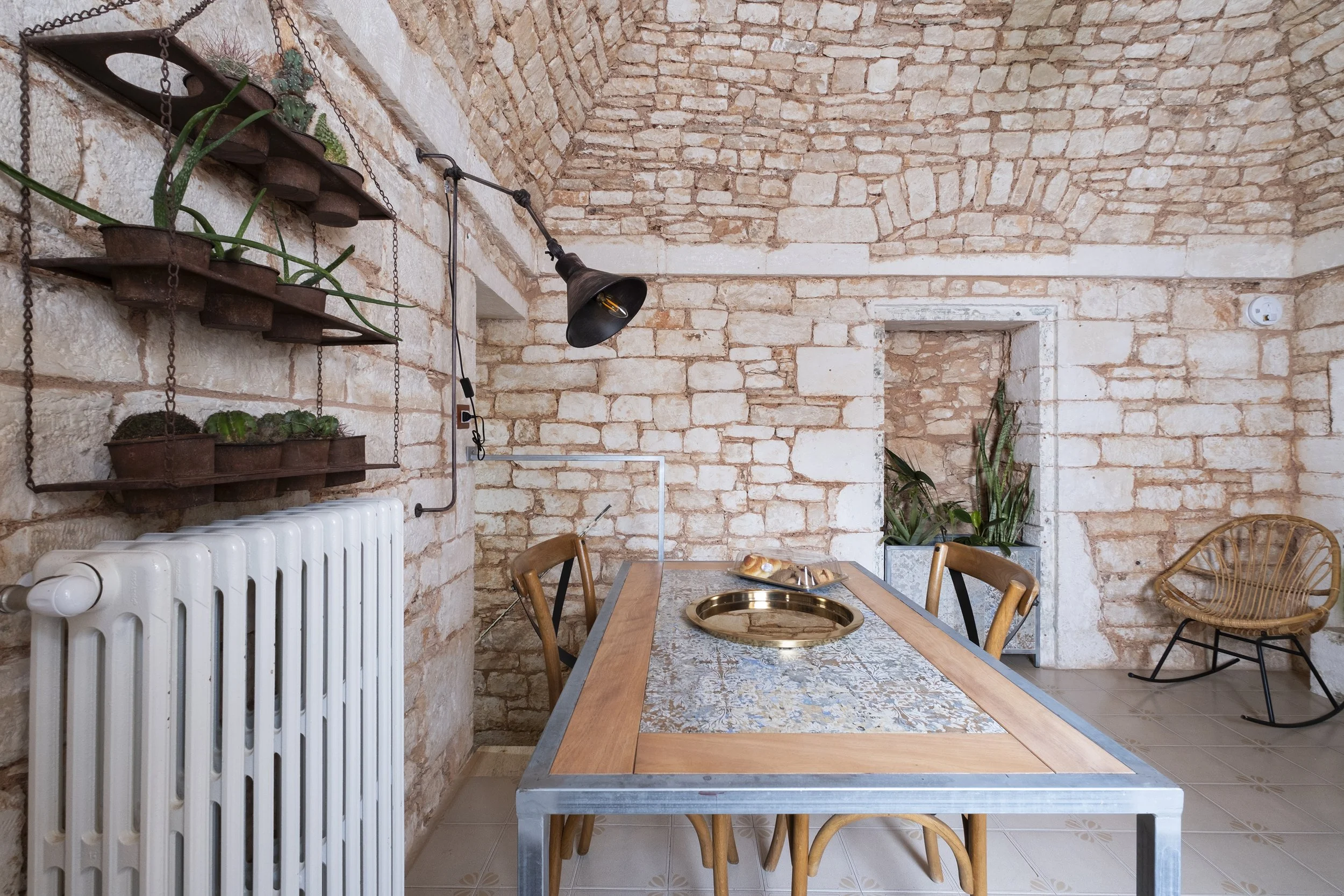 Dining room with exposed brick walls, a rectangular table with a metal frame and wooden inlay top, four chairs, a white radiator, hanging plant shelves, potted plants, and a rattan rocking chair.
