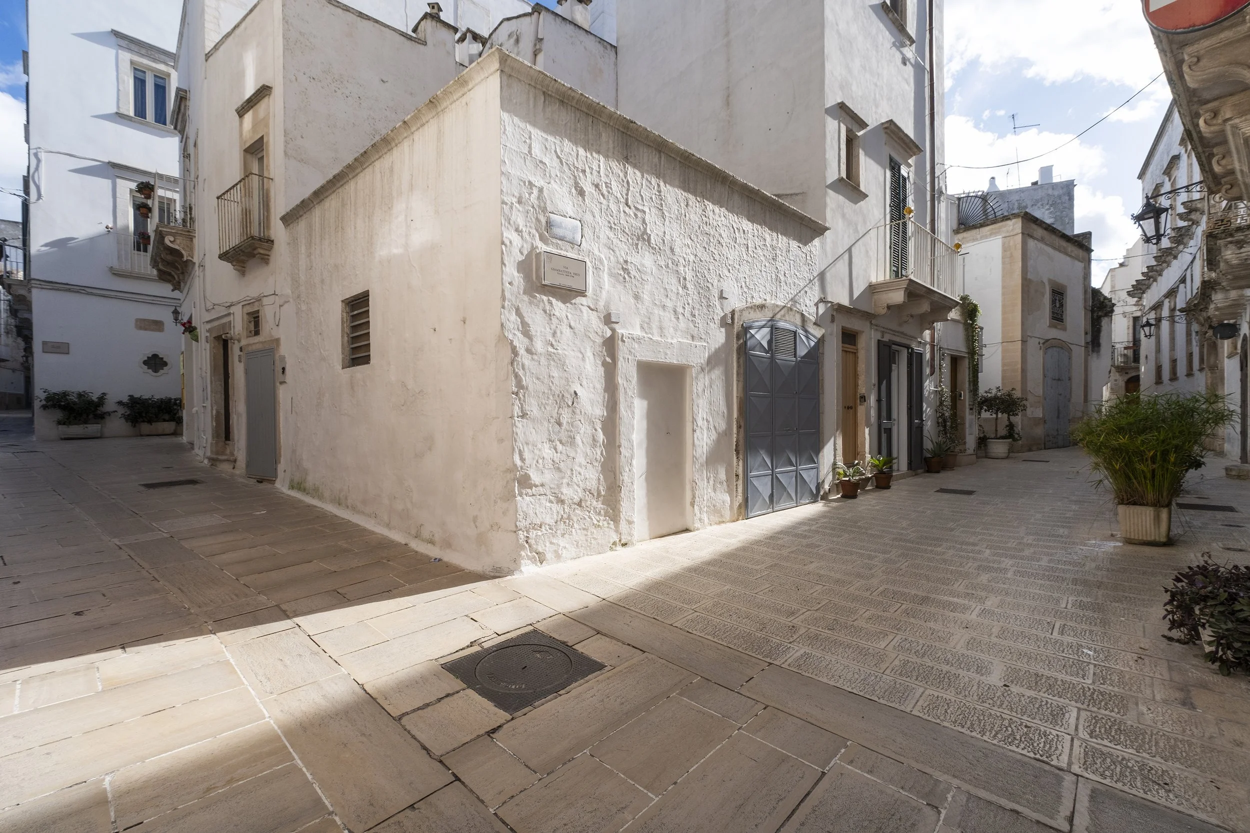 A narrow European street with white and light-colored buildings, potted plants, and a metal gate, under a partly cloudy sky.