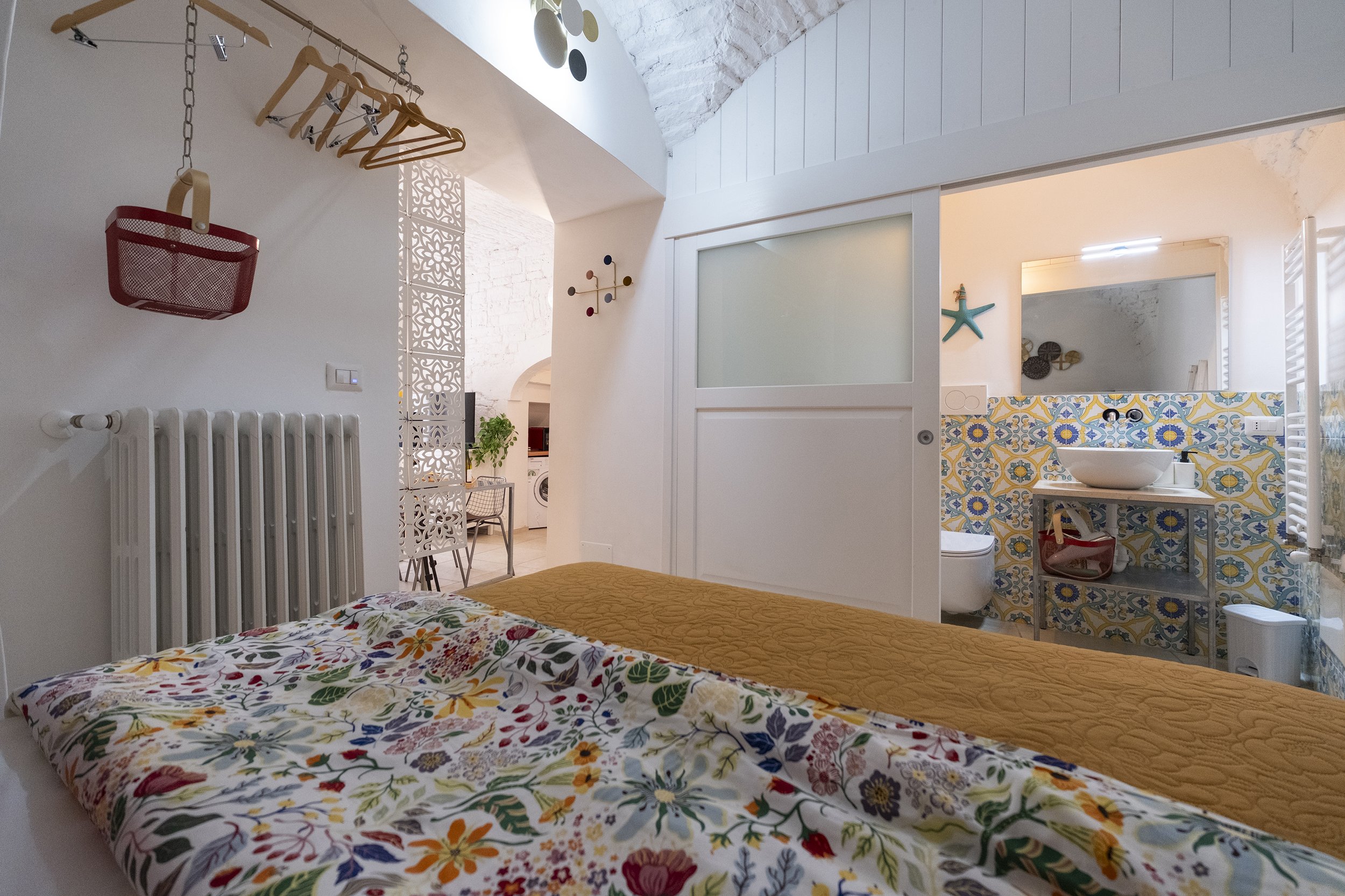 Bedroom with a floral bedspread, a bathroom with colorful tile walls, a sink, and a mirror, separated by a sliding door, in a cozy white-walled apartment.