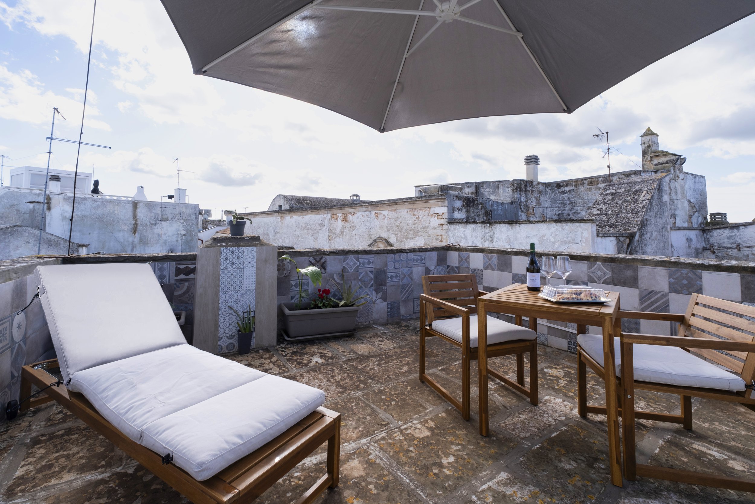 Rooftop terrace with wooden outdoor furniture, a sun lounger, an umbrella, and potted plants, overlooking old stone buildings with weathered roofs.