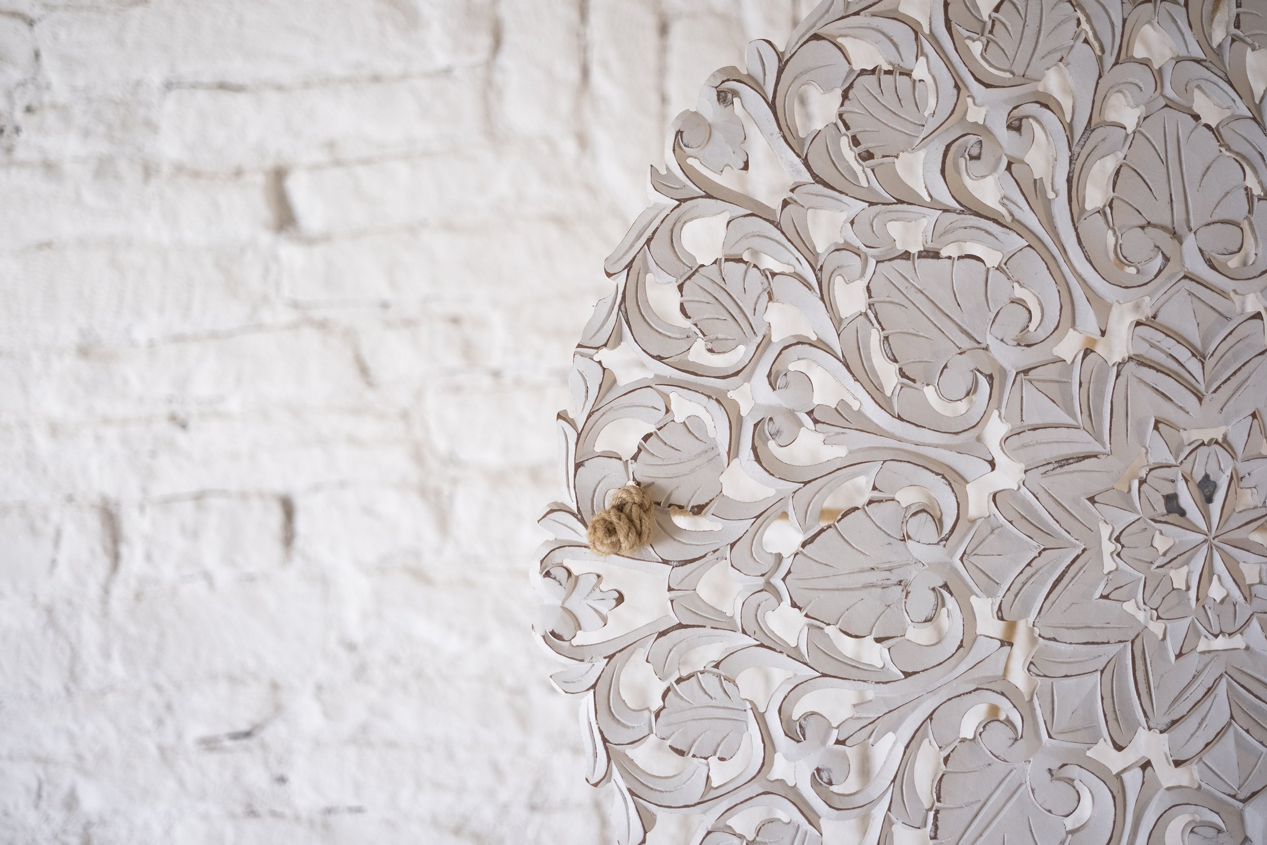 Close-up of a decorative, white carved wooden lampshade with intricate floral and swirl patterns, hanging against a white brick wall.