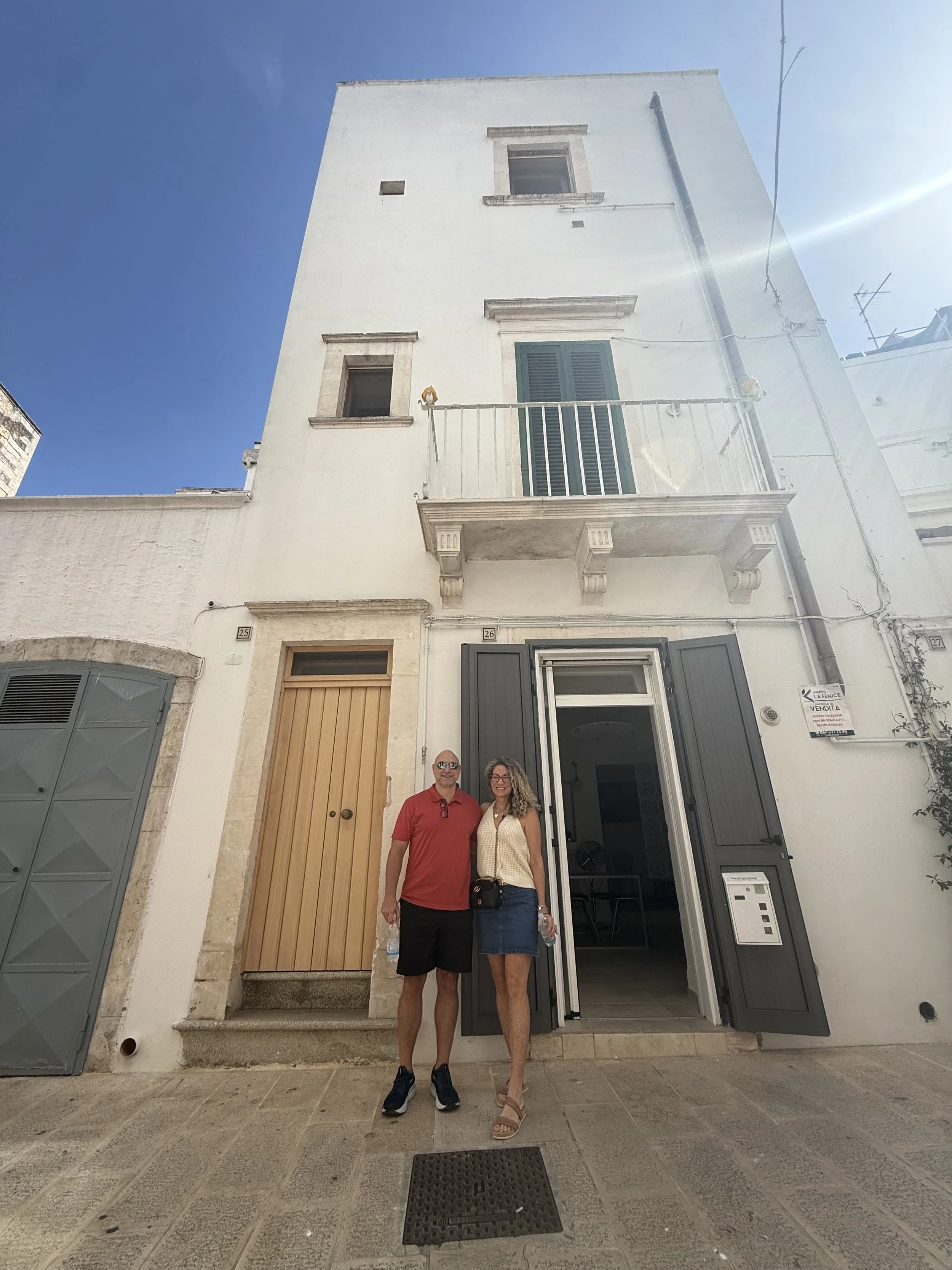 A man and woman standing in front of a white multi-story building with open black shutters, a balcony, and a wooden door, under a clear blue sky.