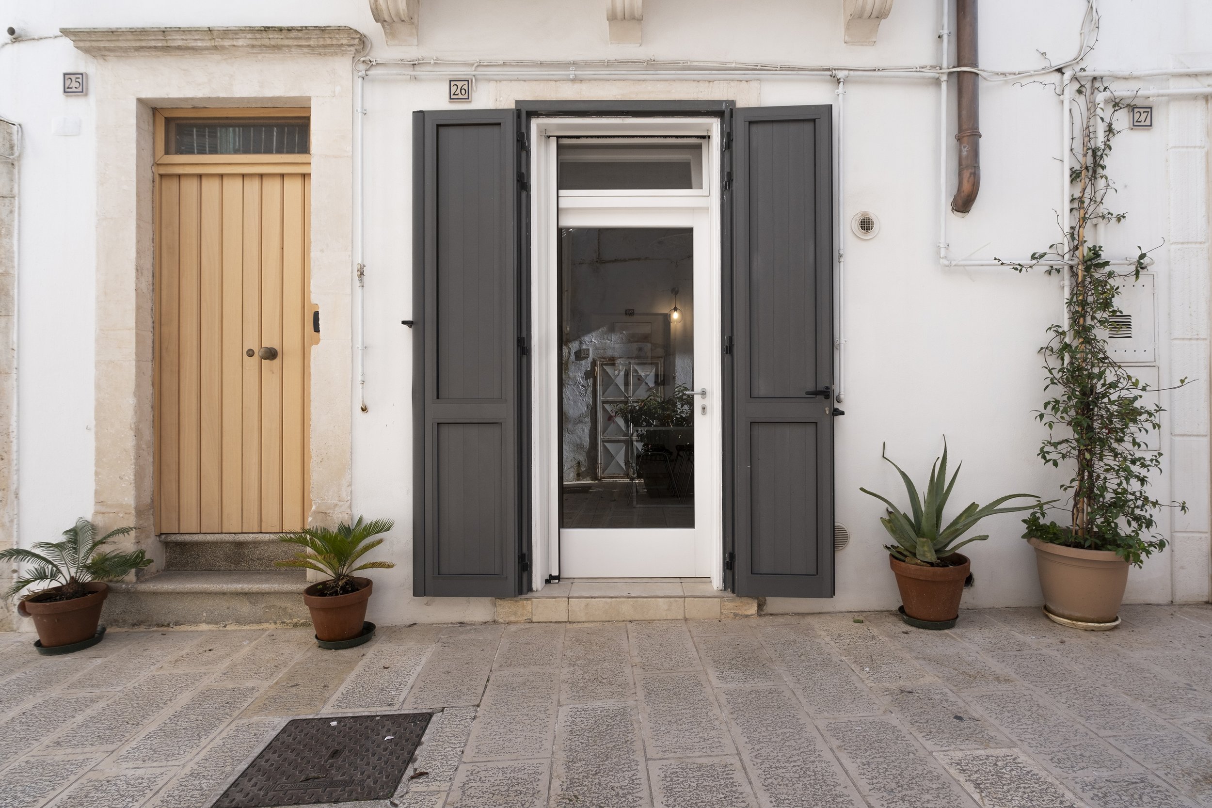 Front entrance of a building with a glass door and black shutters, surrounded by potted plants on a stone-paved sidewalk.