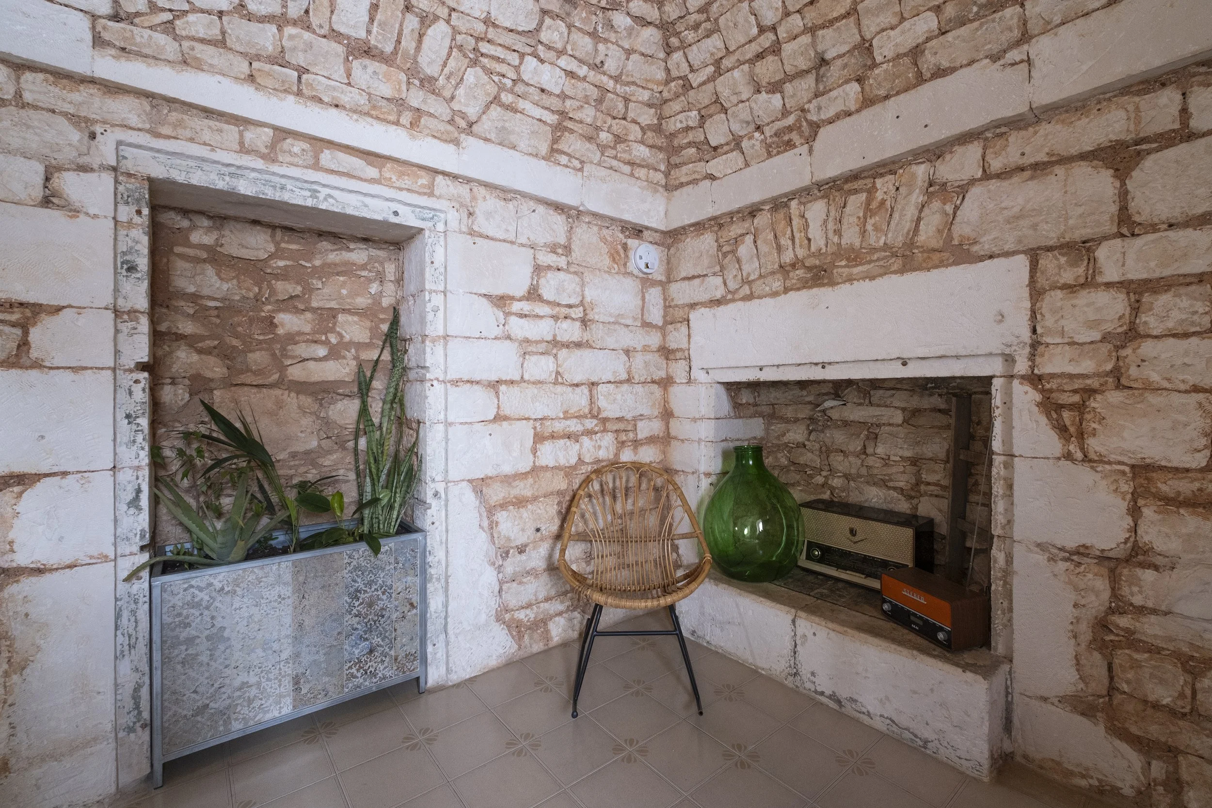 A rustic room with exposed brick walls, a window with potted plants, a wicker chair, and vintage decor including a large green vase and old radios.