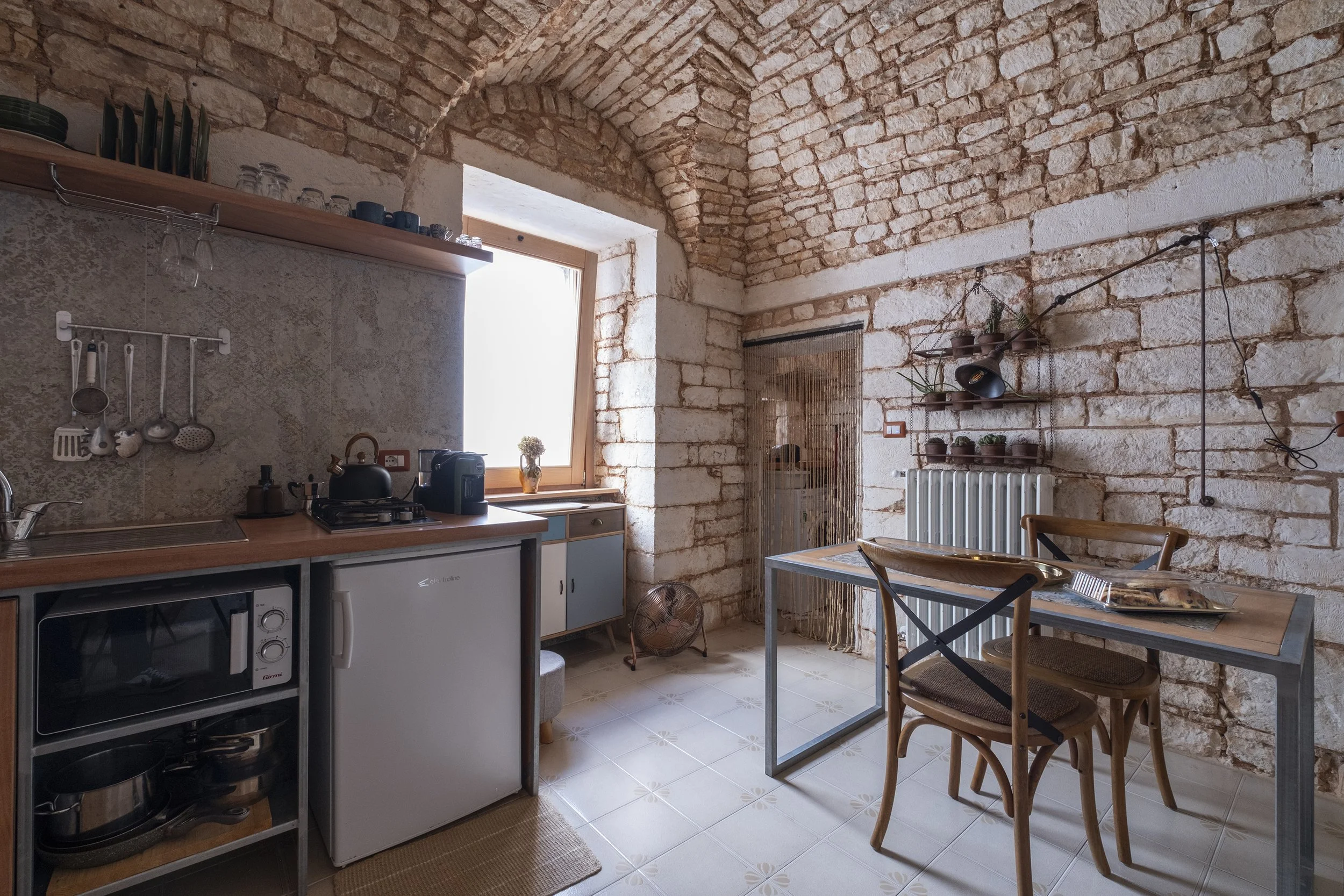 Interior view of a rustic kitchen with exposed brick walls and vaulted ceiling, featuring a small table with chairs, a radiator, and kitchen appliances including a microwave, mini fridge, and stove, with shelves holding dishes and plants.