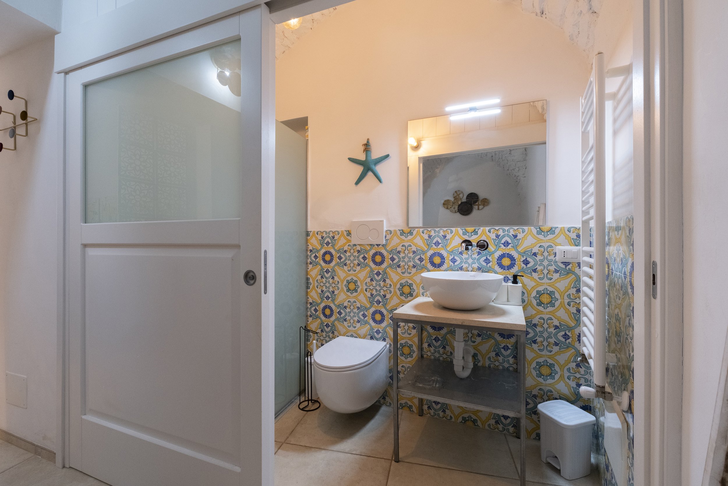 Small bathroom with a colorful tiled wall, wall-mounted toilet, sink on a small table, shower with frosted glass door, and a decorative starfish on the wall.