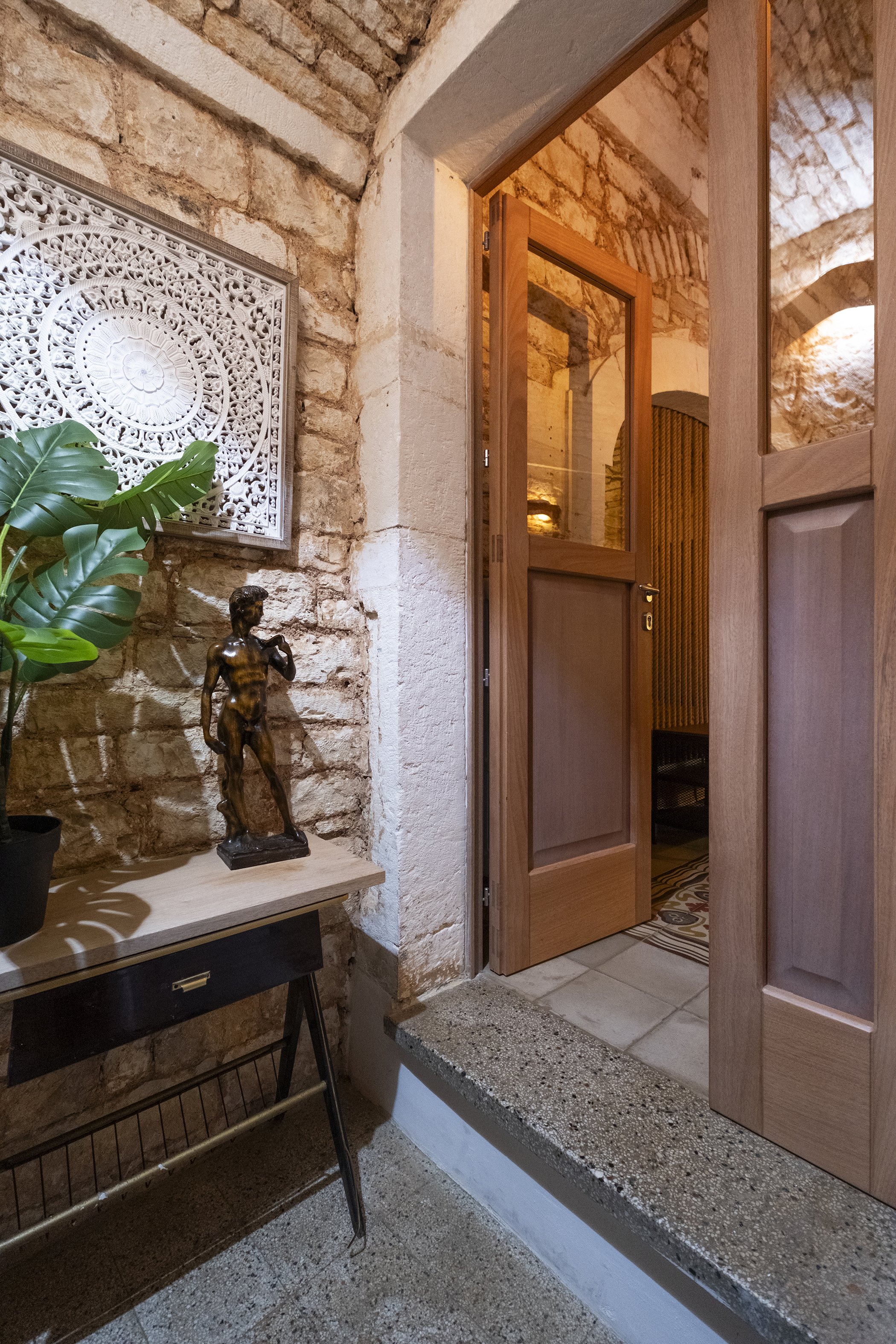 An interior shot of a rustic entryway with exposed brick walls, wooden door, large greenery plant, a small table with a bronze sculpture of a woman, and decorative wall art.