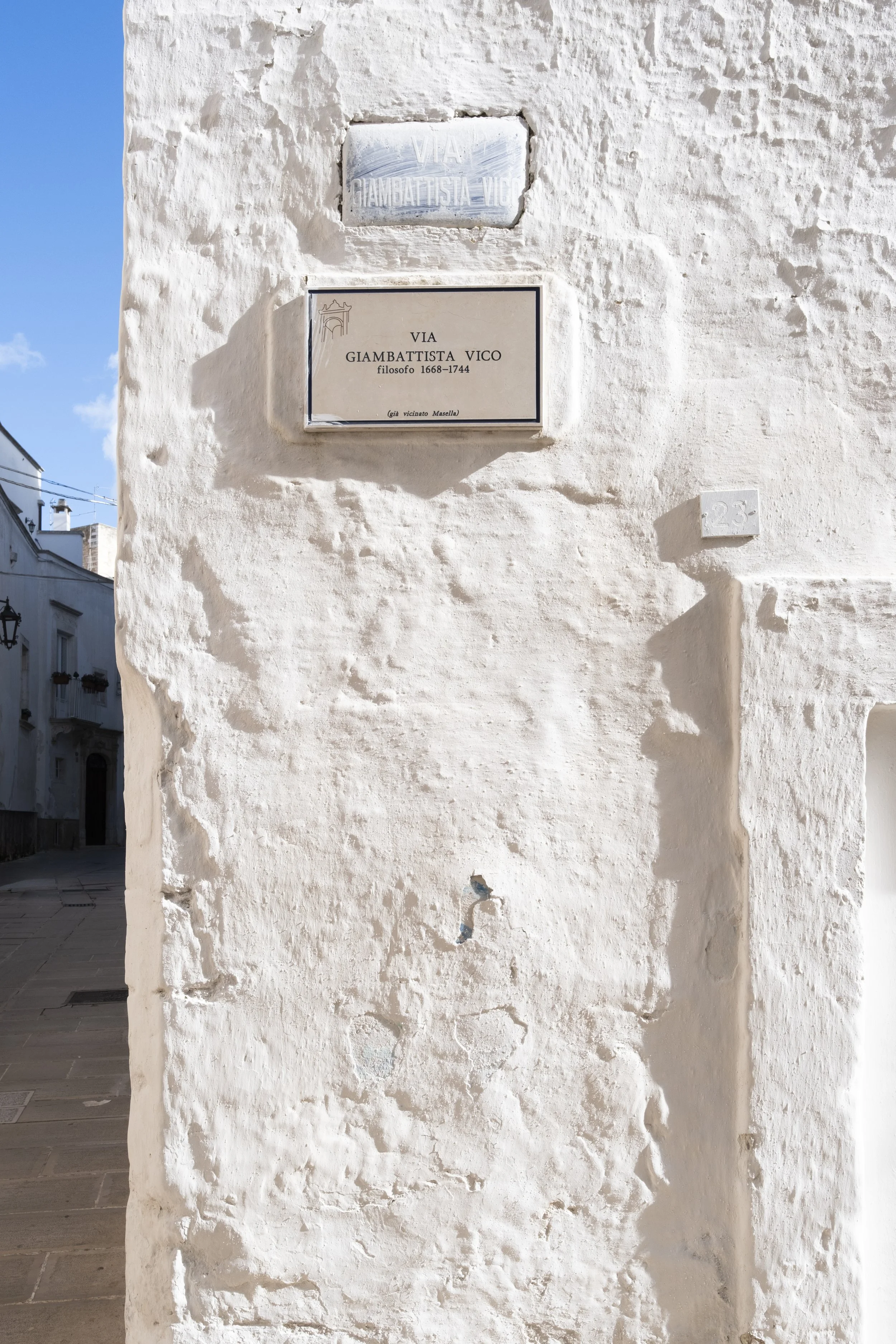 A white stucco wall with street signs reading 'Via Giambattista Vico' and a small plaque with the number 23 on it. In the background, part of a narrow street with similar white buildings.