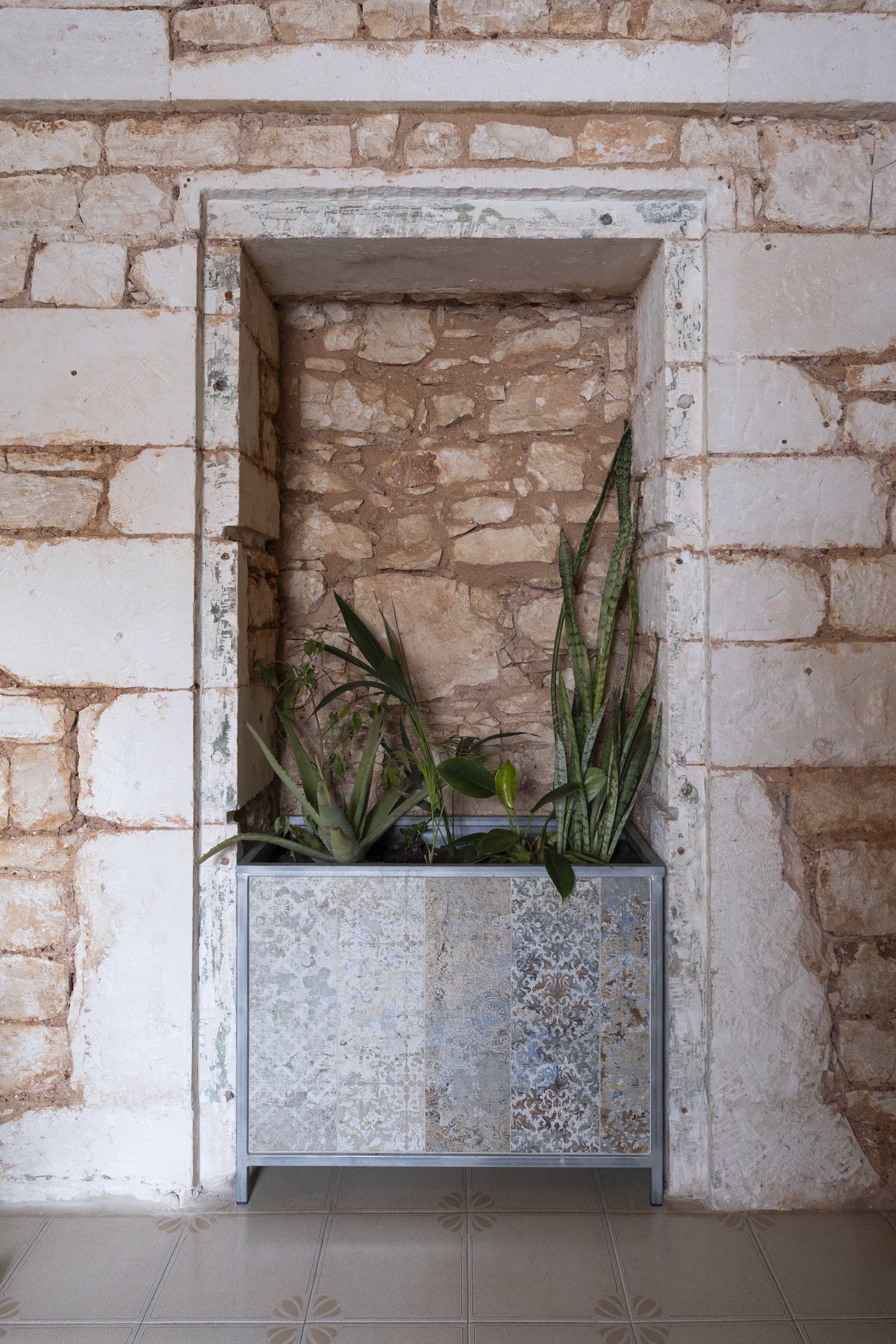 A decorative planter box with a patterned tile design on the front, containing green plants, placed against a textured brick wall with a recessed niche in the wall.